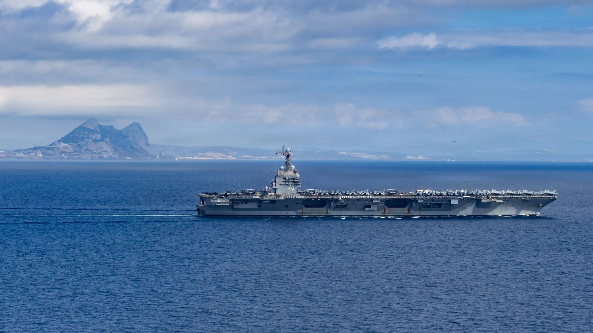 A large aircraft carrier sailing in blue ocean under a partly cloudy sky with mountainous coastline in the distance.