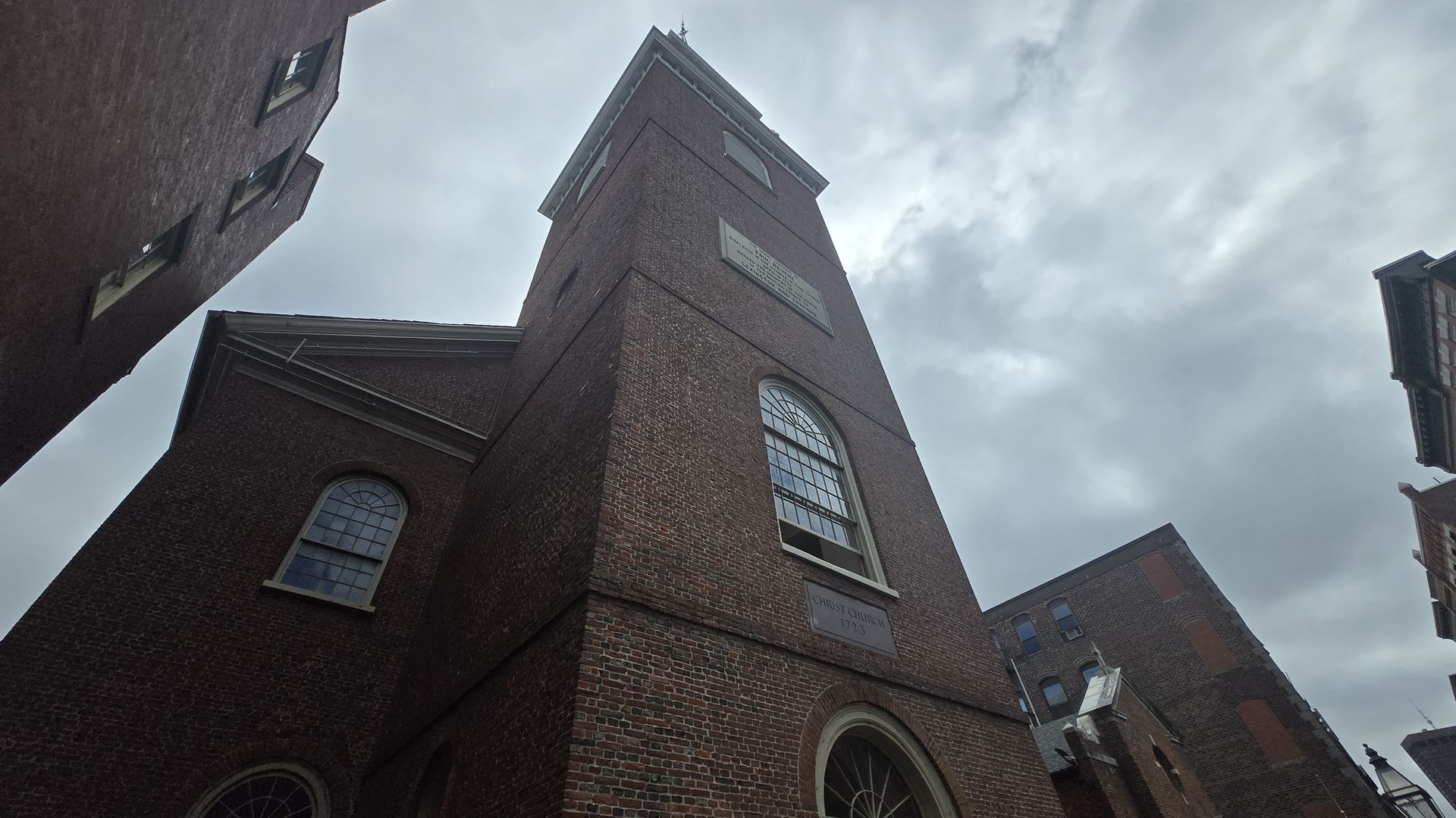 Tall brick tower of Christ Church with large arched windows and plaque, surrounded by other brick buildings under a cloudy sky.