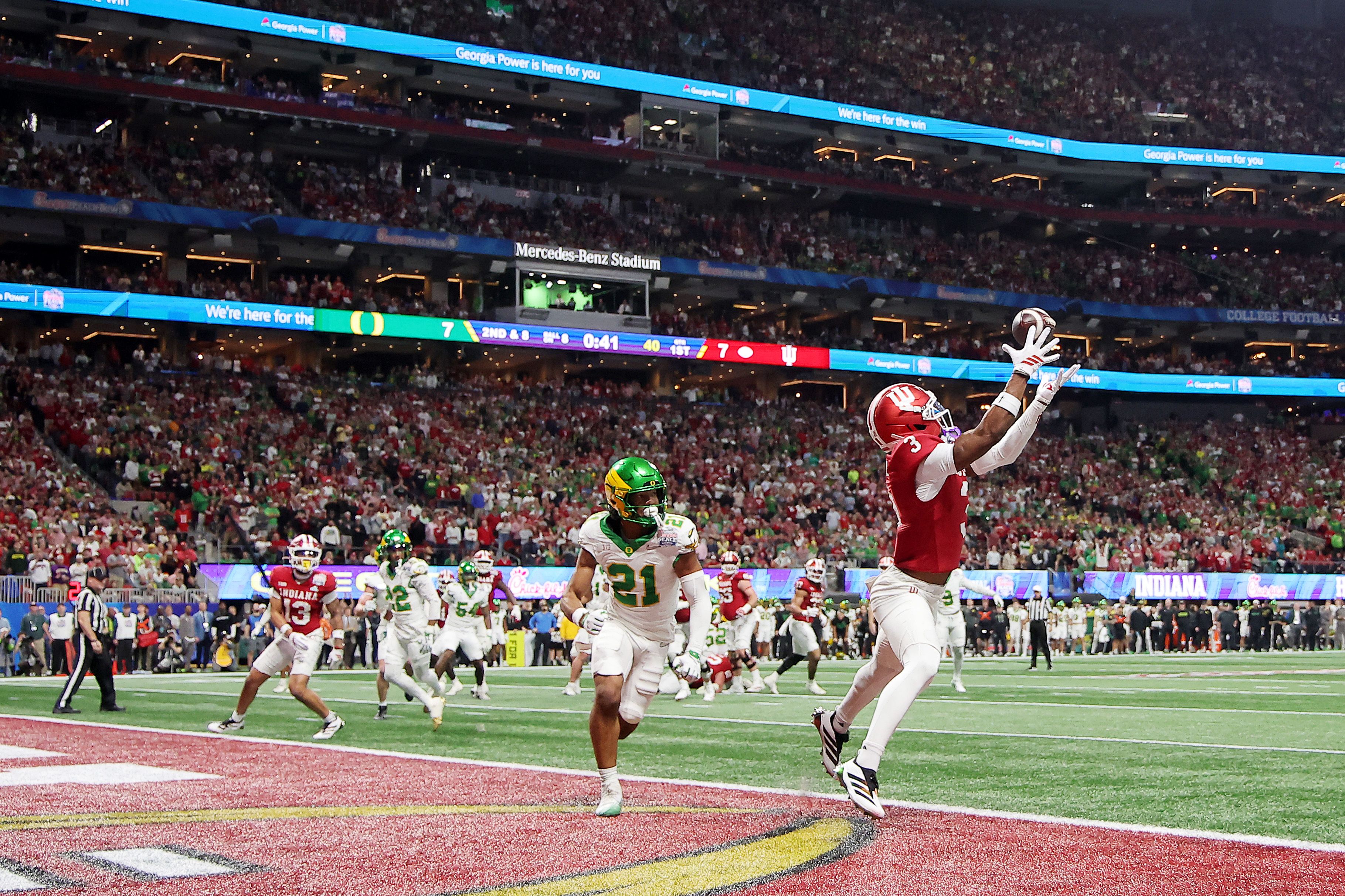 Indiana player in red leaps to catch a football at the edge of the end zone, as a green-and-yellow defender pursues, with a packed stadium in the background.
