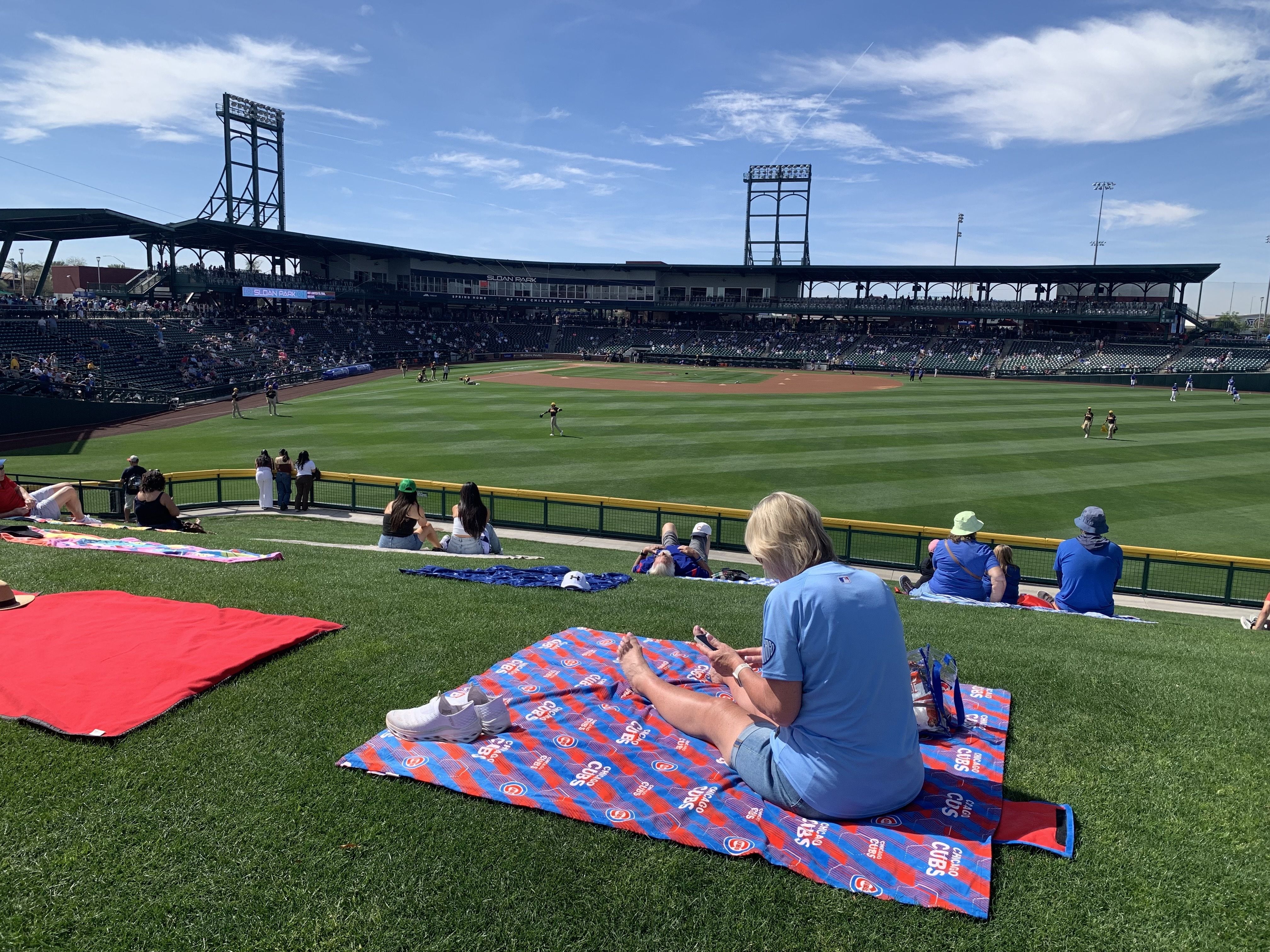 People sit on a grassy hill covered with Chicago Cubs blankets, watching a baseball game at Sloan Park under a sunny blue sky with scattered white clouds.