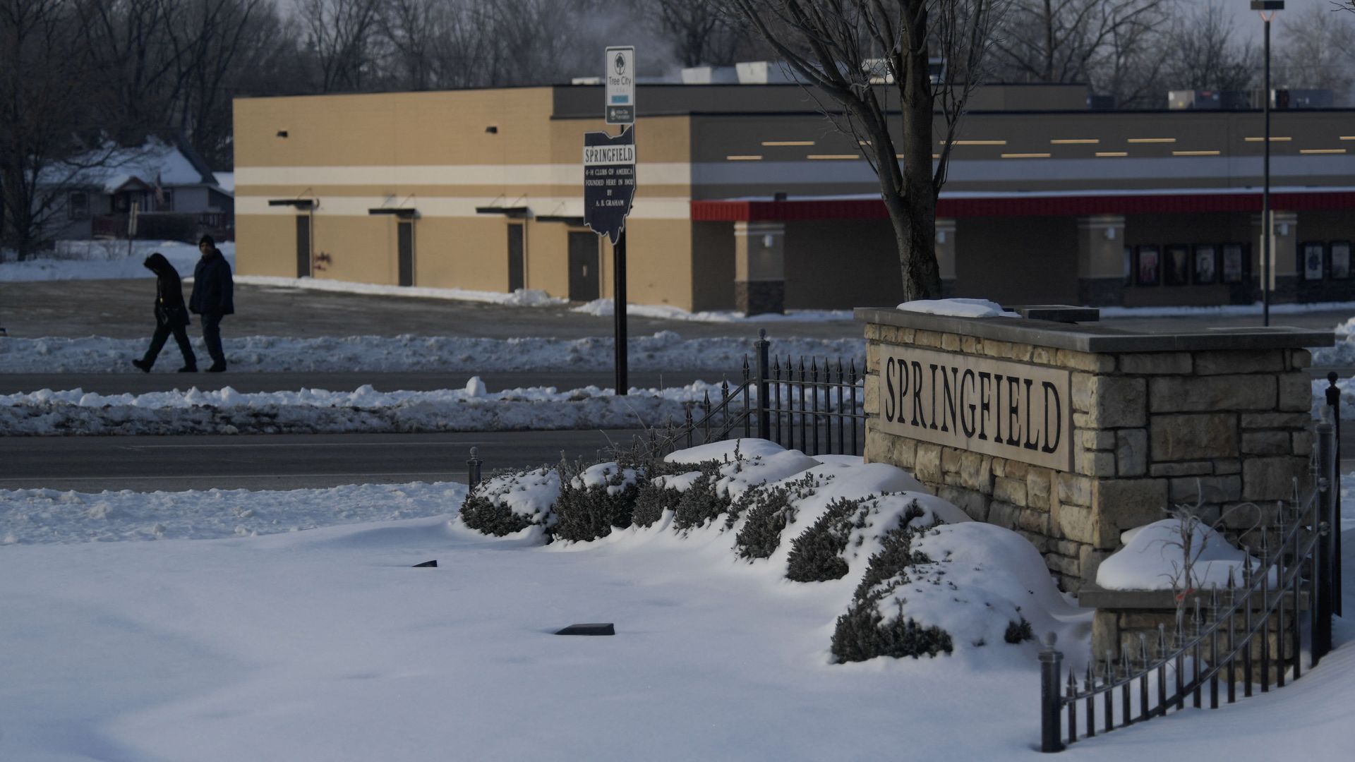 Snow-covered ground with bushes and a stone sign reading "SPRINGFIELD" near a barren tree. Two people walk along a snowy road with a beige and red building in the background.
