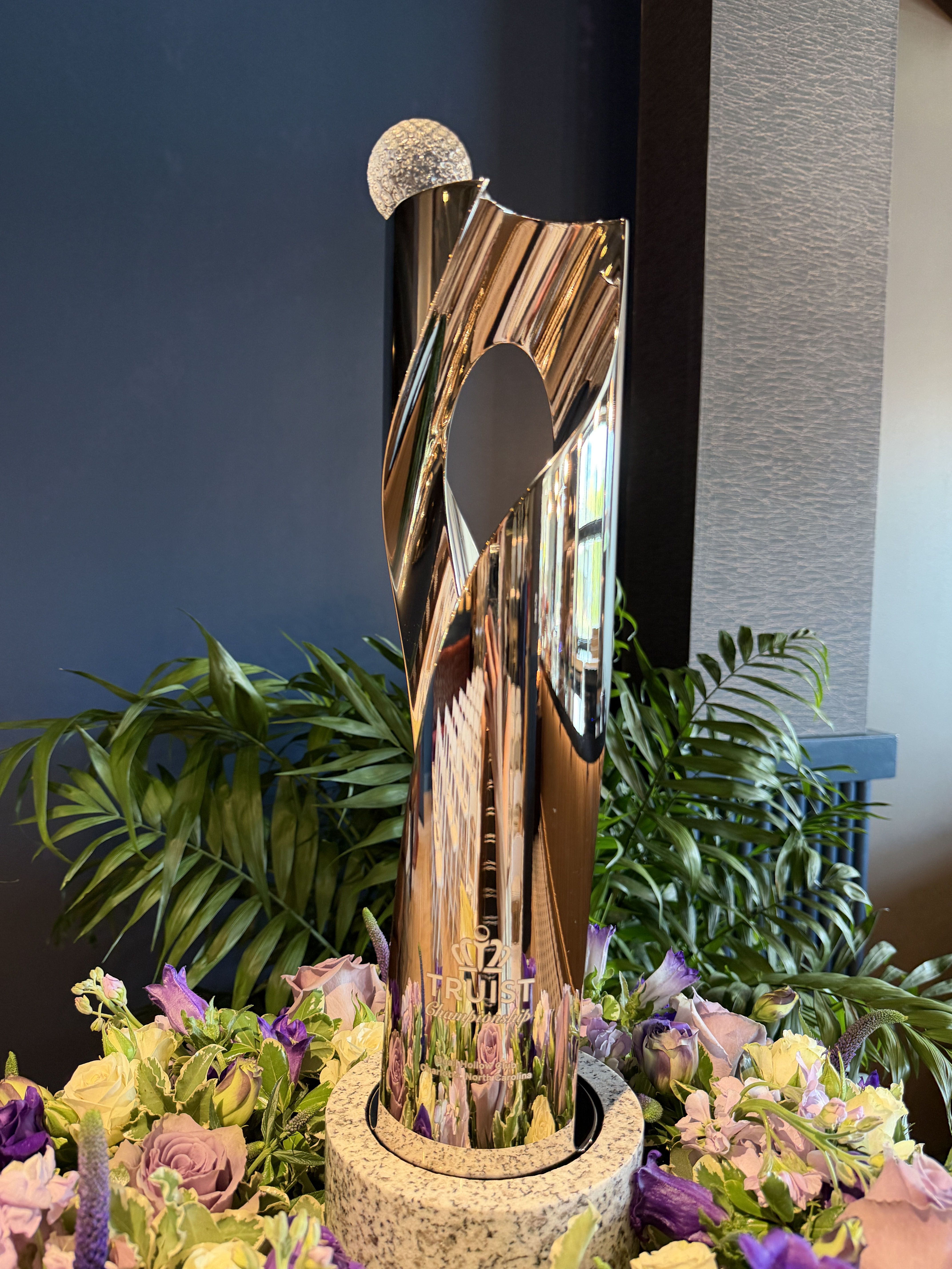 Shiny abstract chrome sculpture with curved shapes stands on a speckled circular base, topped by a crystal ball. Surrounded by pastel roses and purple flowers with green plants against blue and gray walls.