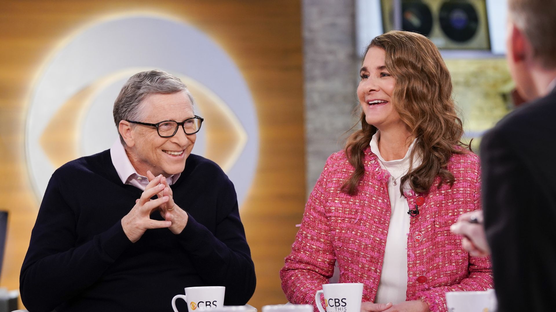 Bill Gates and Melinda Gates laugh on the set of CBS this morning, with the CBS symbol in the background.