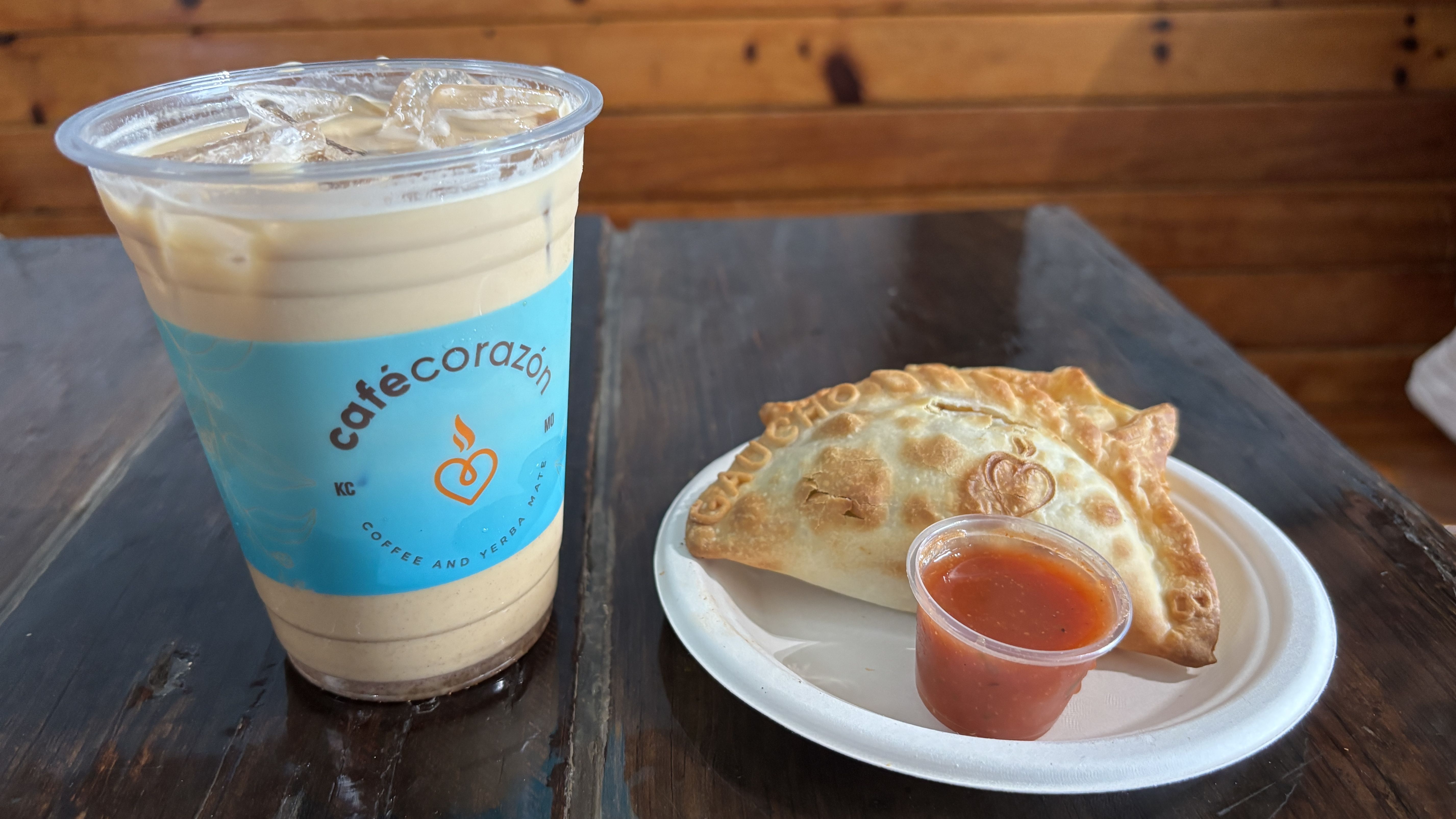 Iced coffee in a clear cup with a blue sleeve sits on a dark wooden table next to a plate with a baked pastry and a small cup of red sauce, against a wooden wall.