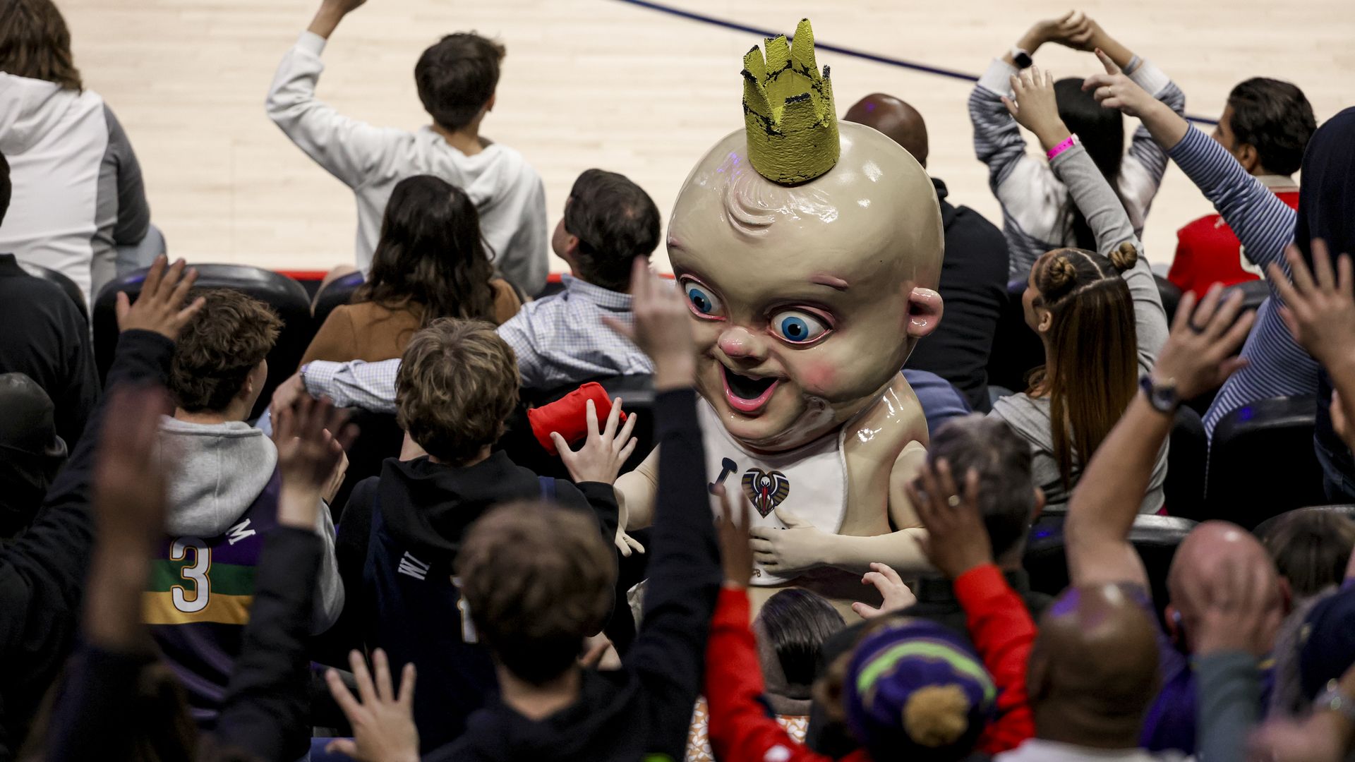 A large NBA mascot dressed as an oversized, crowned baby with a bib miingles amongst the audience during a basketball game.