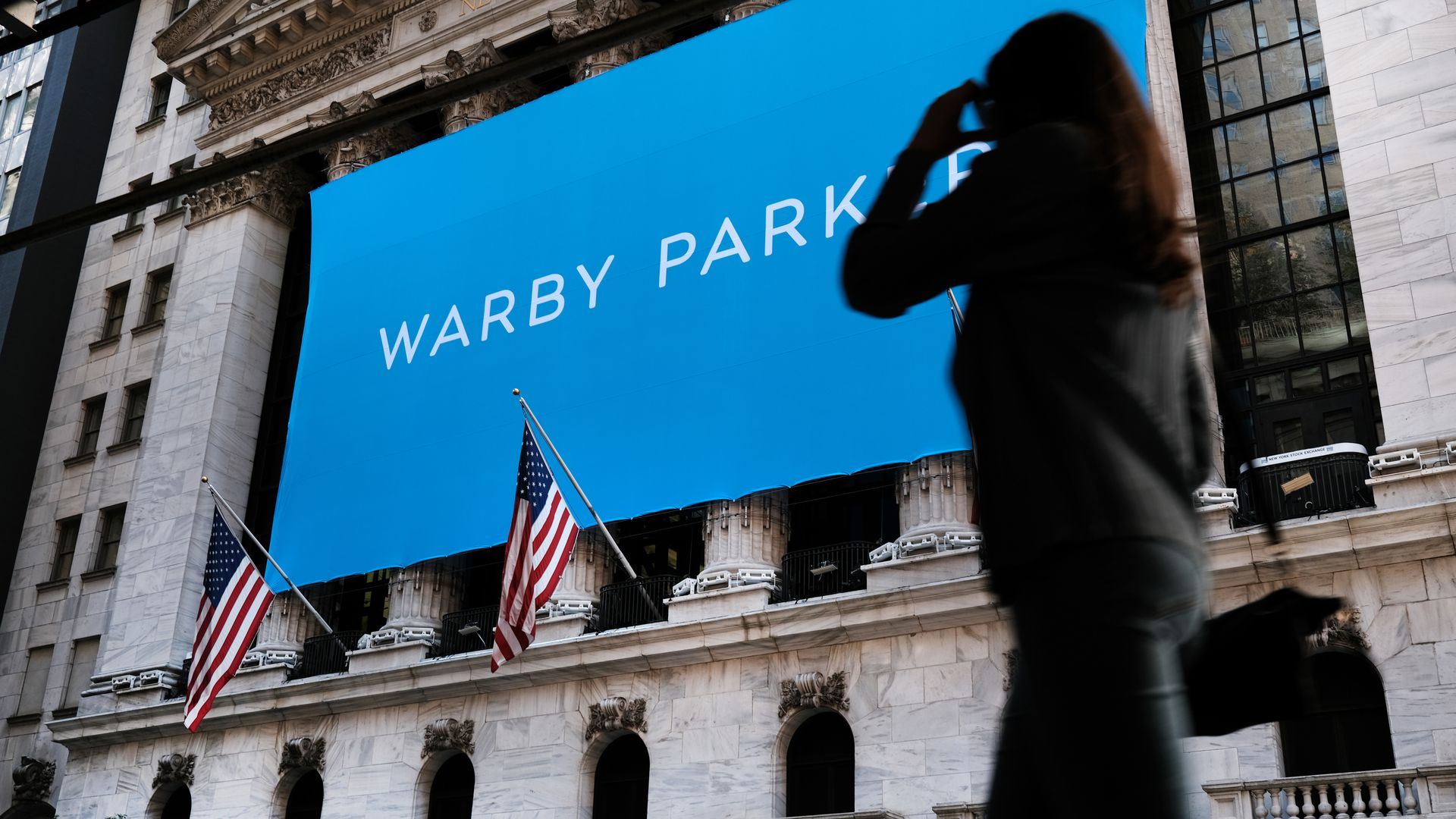 Photo of a person walking in front of a building with a huge screen that says "Warby Parker"