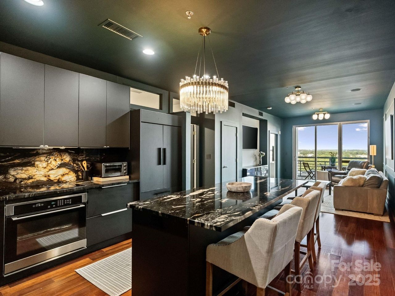 Modern black kitchen with marble countertops, black cabinets, chandelier lighting, and wooden floor leading to a living area with gray sofa, chairs, and large windows overlooking greenery.