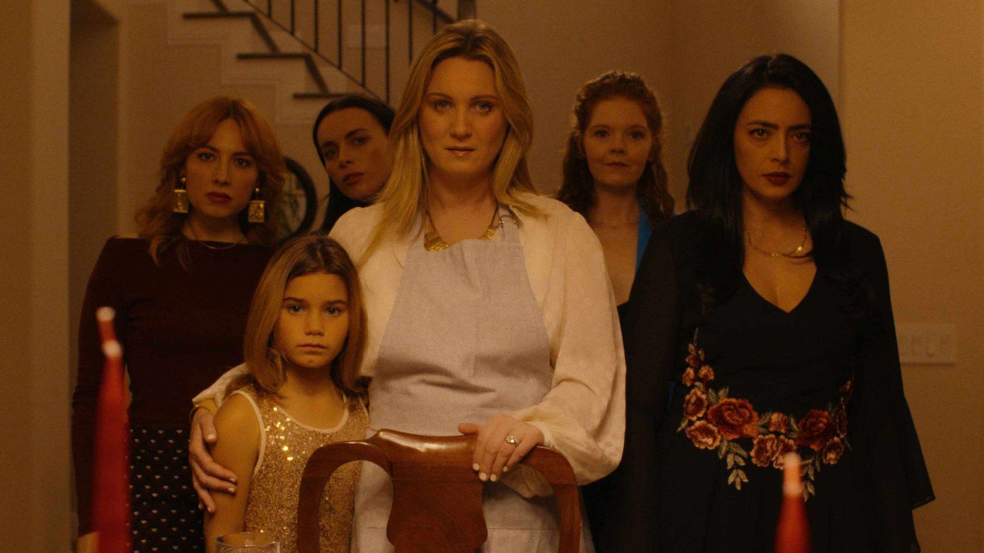 Five women and a young girl stand in a warmly lit hallway. Center is a blonde woman in a pale blouse and blue apron, gripping a wooden chair; others flank her, with a red candle nearby.