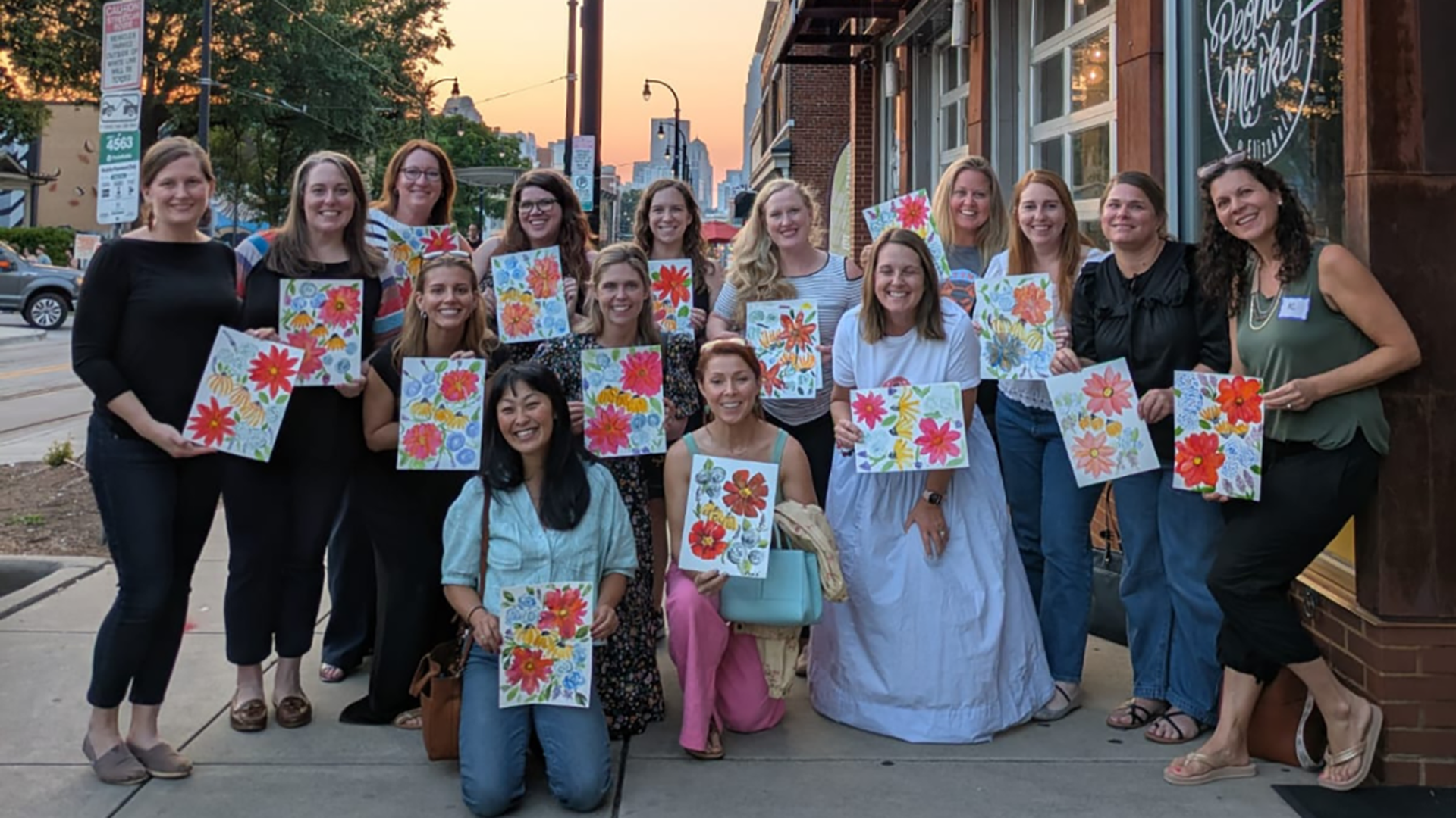 Group of 15 women outdoors at sunset, smiling and holding up colorful flower paintings, standing and kneeling on a sidewalk beside a building with large windows and a sign reading "The People's Market."