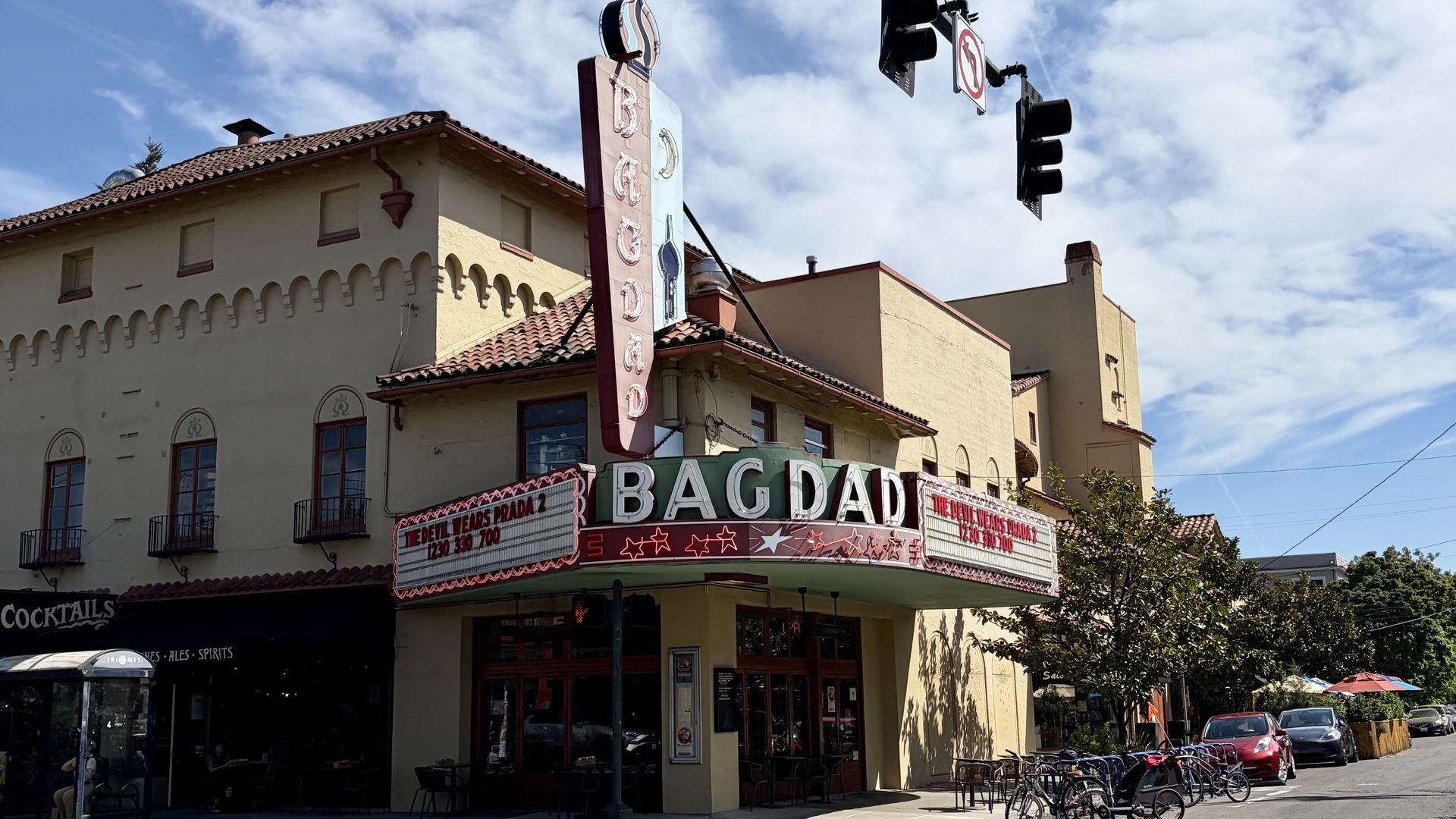 Beige theater building with a red-tiled roof and a tall vertical sign reading "BAGDAD", a red marquee with stars, bicycles parked out front, and a clear blue sky.