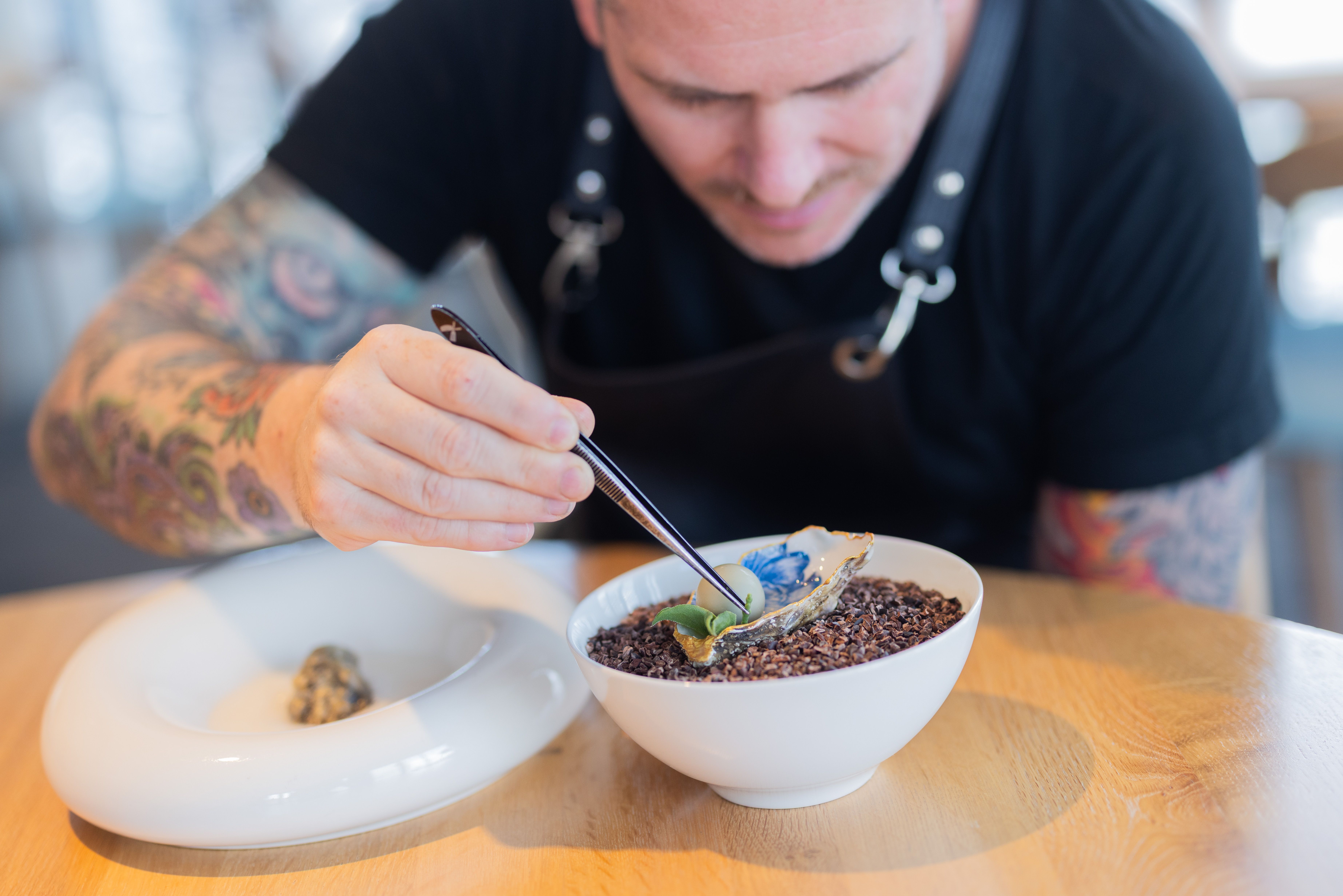 A chef leans over a bowl with a pair of tweezers to delicately place leaves atop what appears to be a bowl of black rice. 