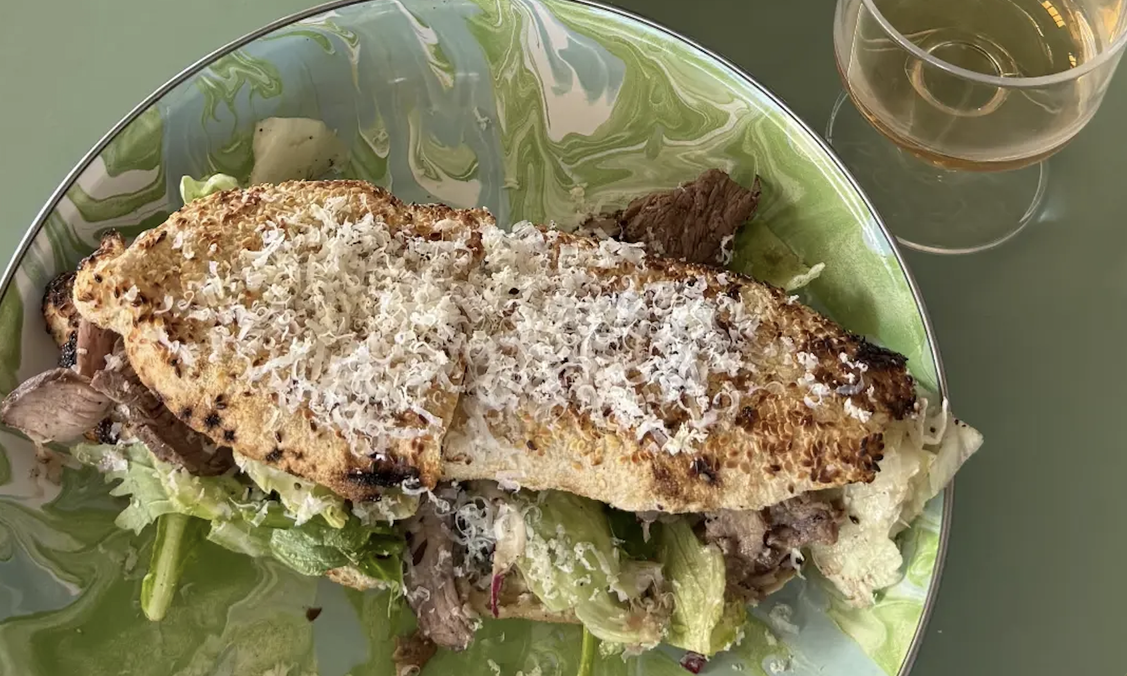Top-down view of a green marbled plate with a long toasted sandwich topped with grated cheese, lettuce and beef; a glass of white wine sits nearby.