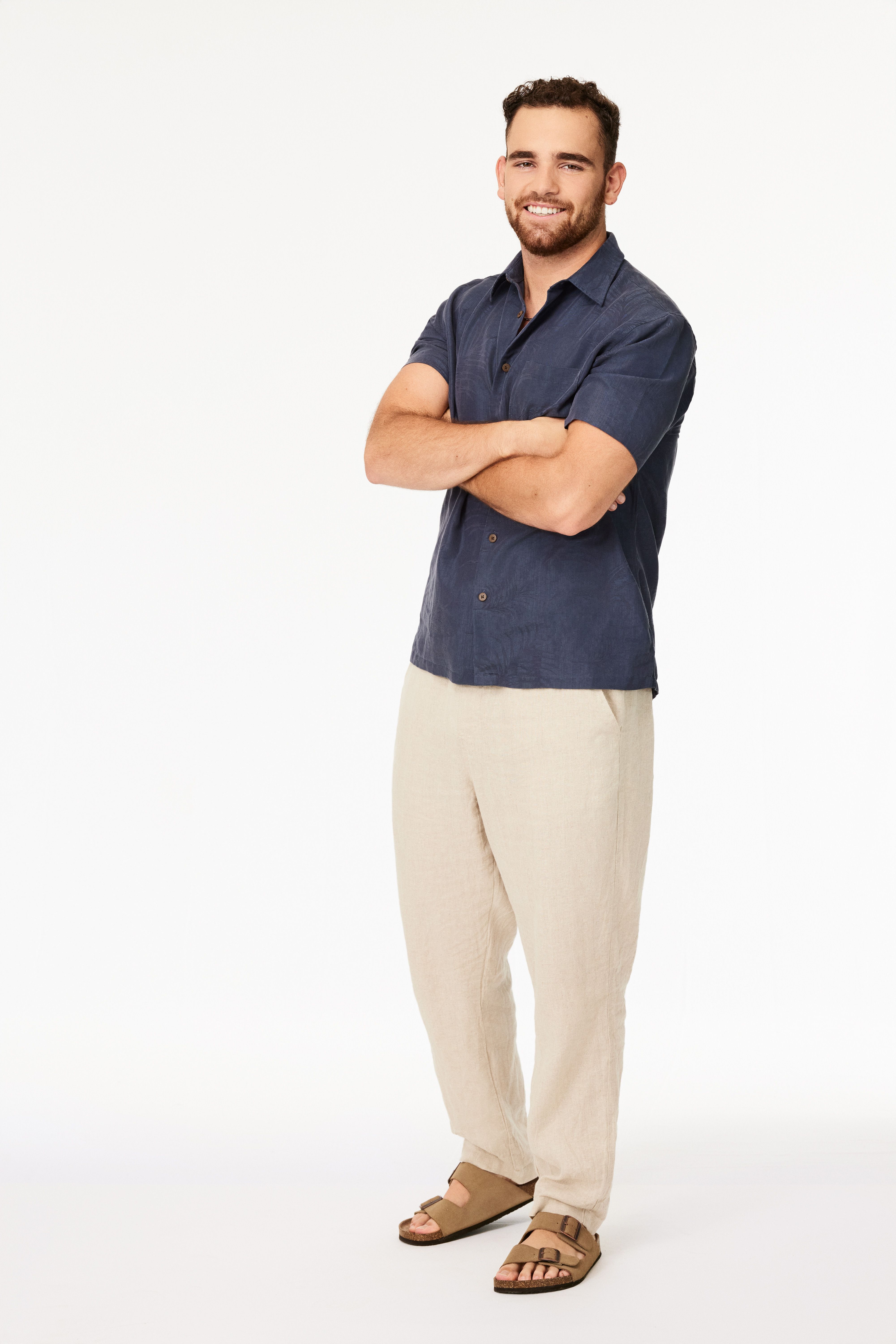Smiling man with short curly hair and beard, wearing a dark blue short-sleeve shirt, beige pants, and brown sandals, standing with arms crossed against a white background.