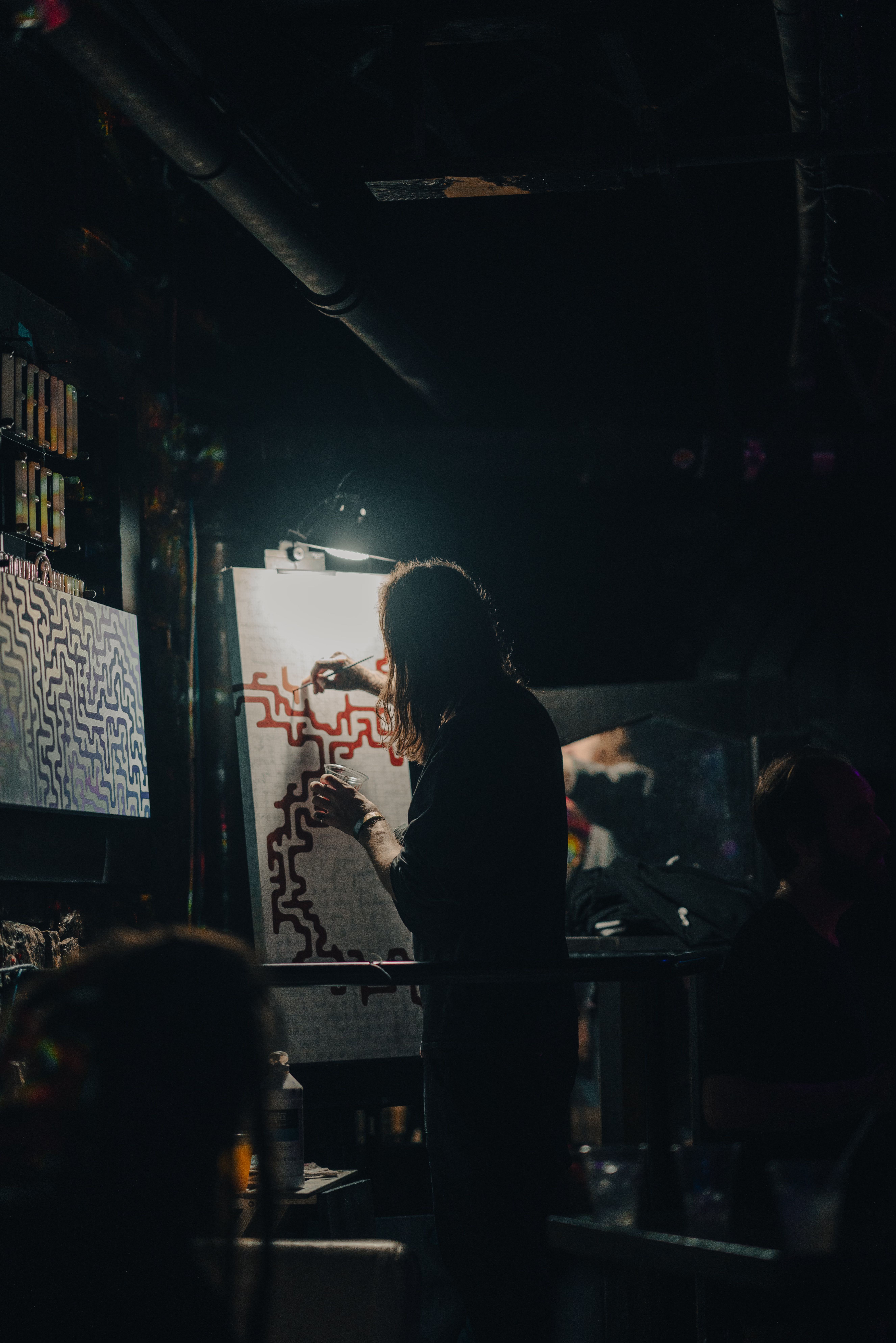 Silhouetted artist with long hair painting red abstract lines on white canvas in dimly lit room, holding a cup, with another artwork featuring blue and purple lines hung nearby.