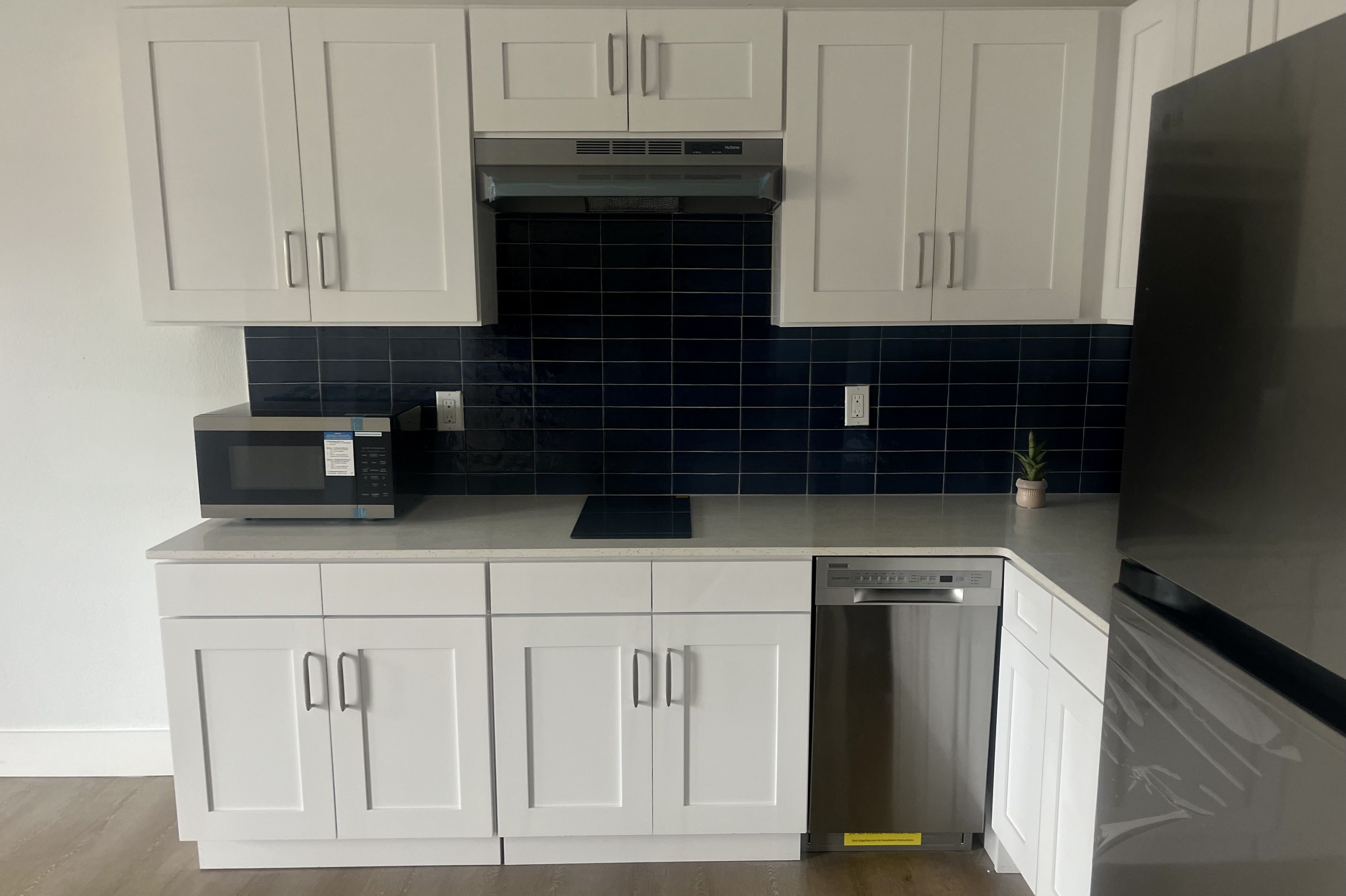 Modern kitchen with white cabinets, dark blue tile backsplash, stainless steel microwave, dishwasher, refrigerator, and a small potted plant on light-colored countertop.