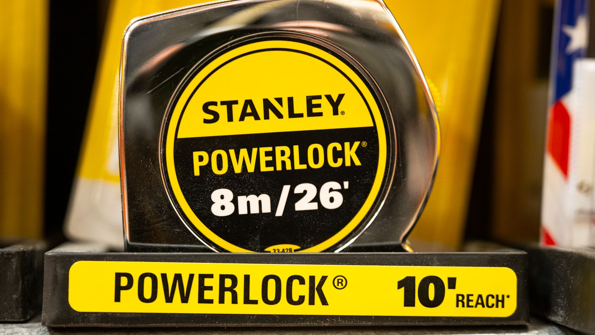 Close-up of a Stanley PowerLock tape measure in a black chrome case, featuring a bright yellow circular label with STANLEY POWERLOCK and 8m/26' markings; yellow background.