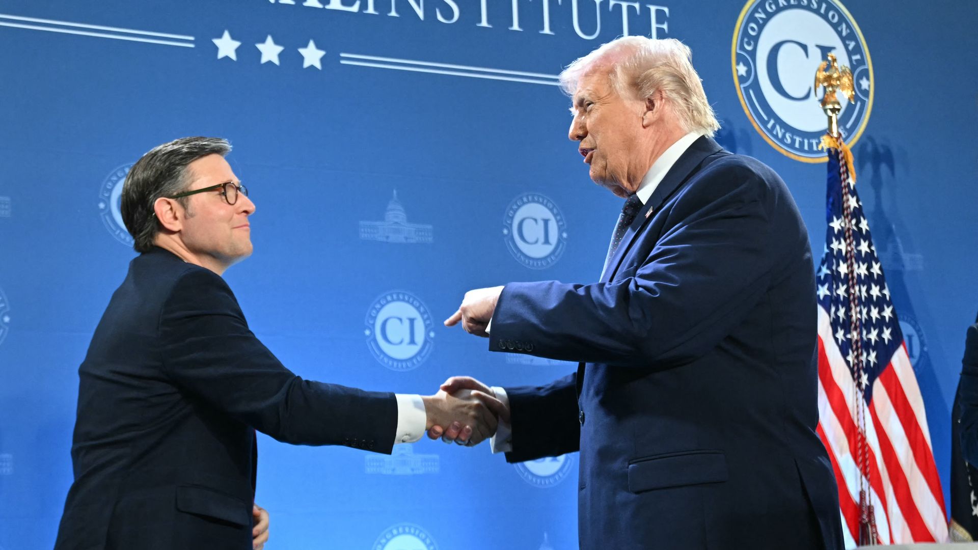 Two men in dark suits shake hands on a blue stage, backdrop filled with circular CI logos; an American flag stands to the right, signaling a formal event setting.