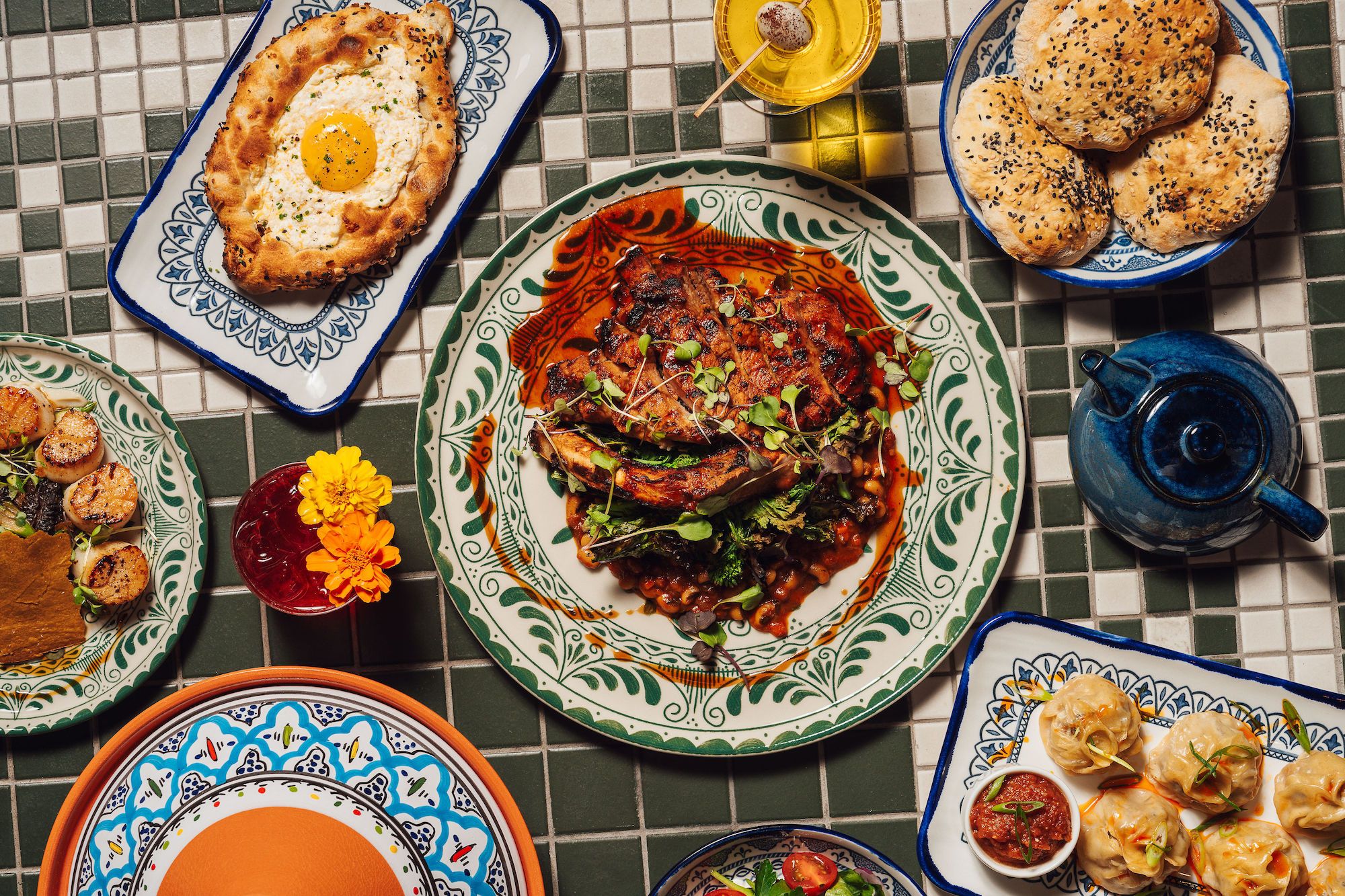 Overhead view of a table with various dishes including grilled meat with sauce on a large decorated plate, bread rolls with black seeds, dumplings with red dipping sauce, a flatbread with egg, grilled scallops, a teapot, and a red drink with yellow flowers.