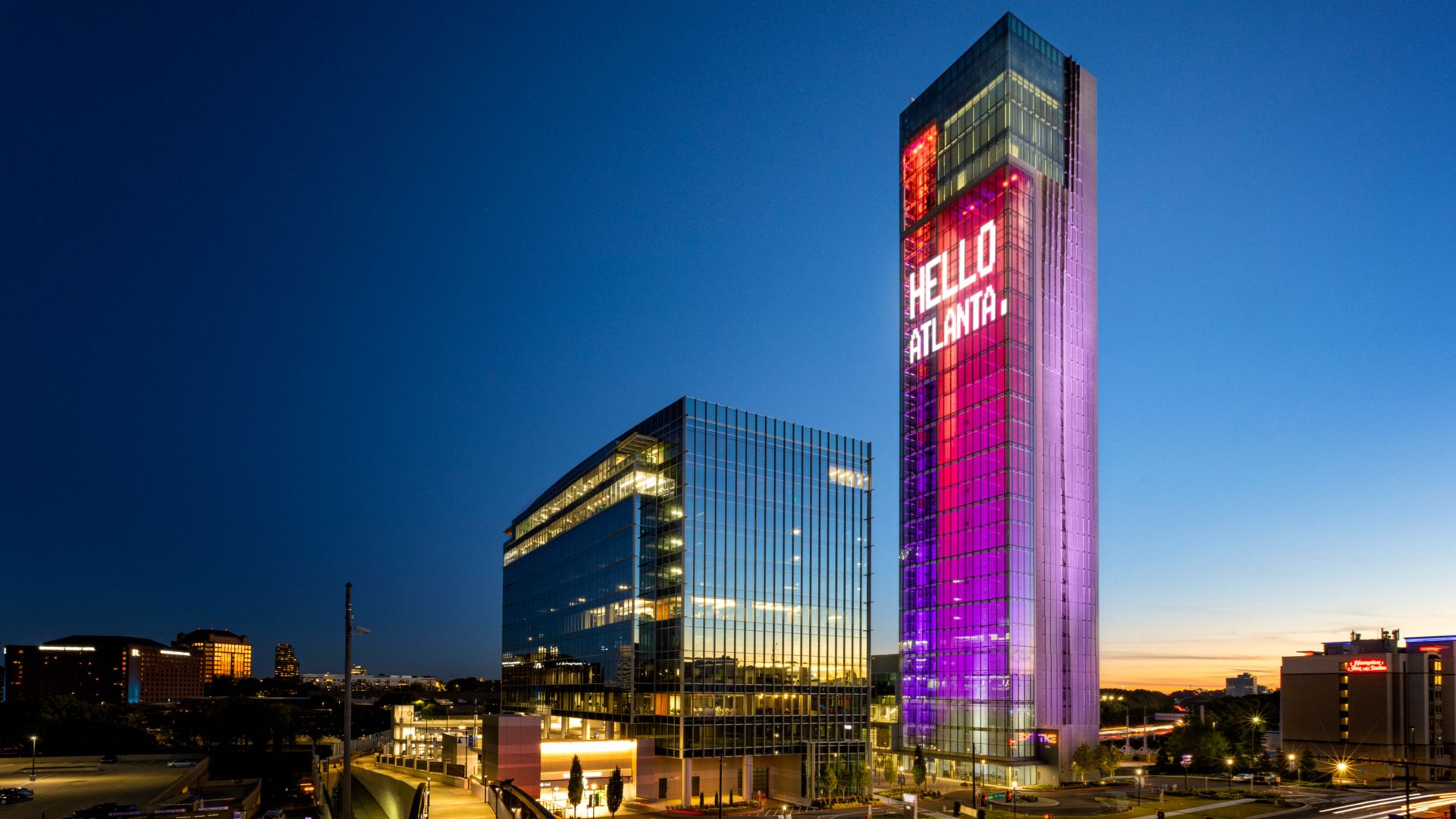 A tall tower illuminated in purple and red beams of light with a large digital sign reading "Hello Atlanta"