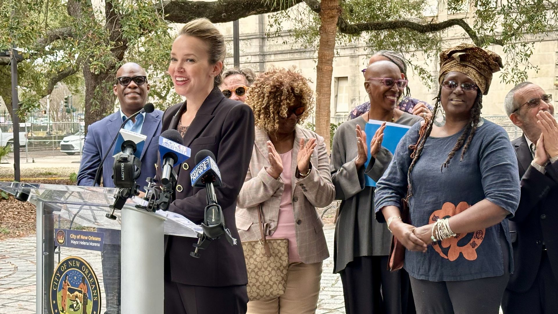 A woman stands at a podium with microphones in front of a City of New Orleans seal, speaking and smiling. Several people behind her are clapping outdoors under trees.