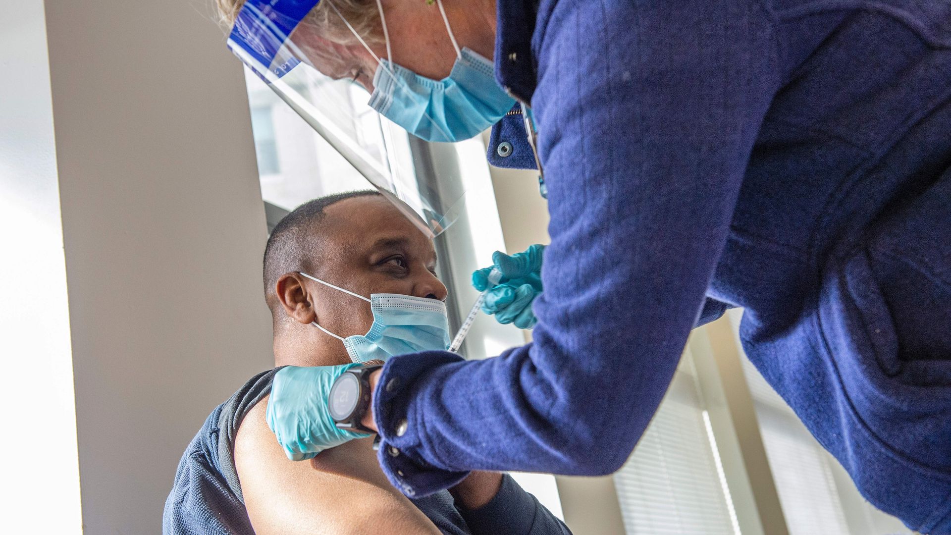Photo of a masked person getting a vaccine shot from a health care worker wearing a face shield