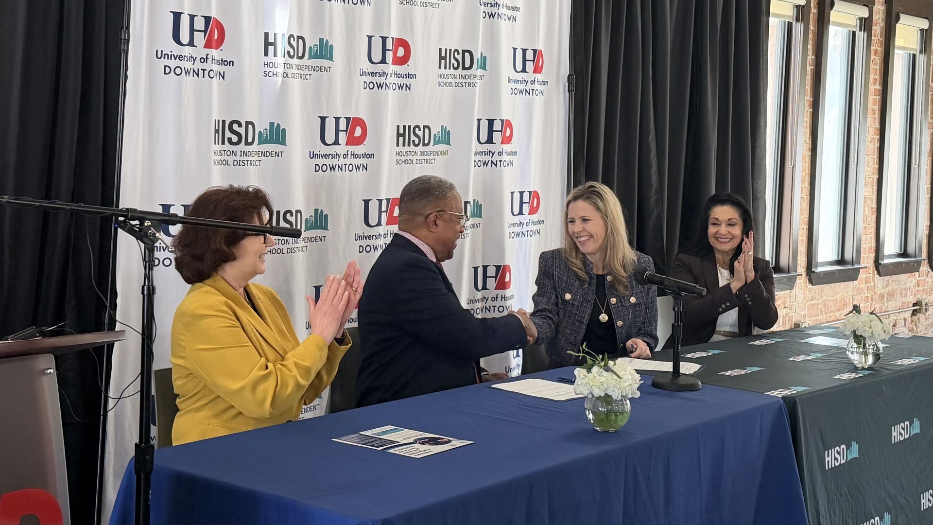 Four people seated at tables with blue and green cloths showing University of Houston Downtown and Houston Independent School District logos, two individuals shaking hands and others clapping in a brick-walled room.
