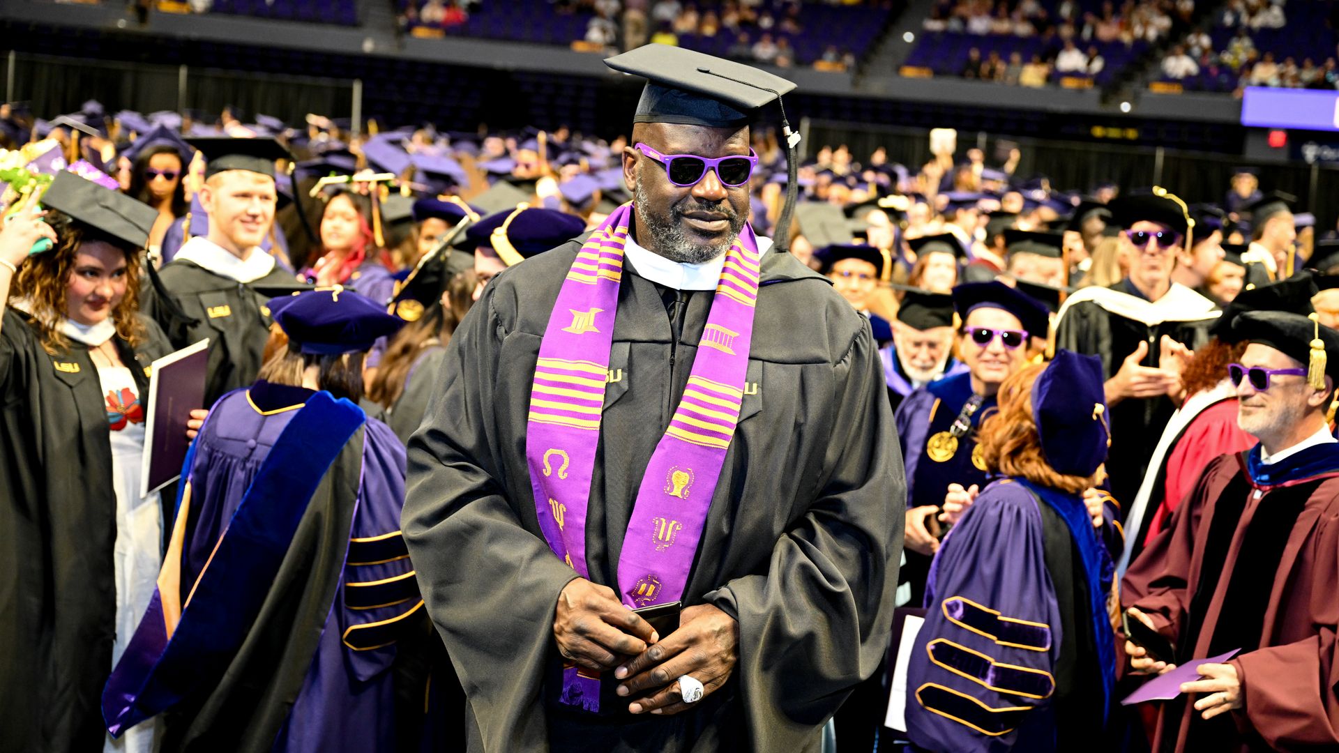 A graduate in a black cap and gown with a purple stole and purple sunglasses walks through a crowded graduation ceremony, surrounded by fellow graduates in purple gowns and a cheering audience.