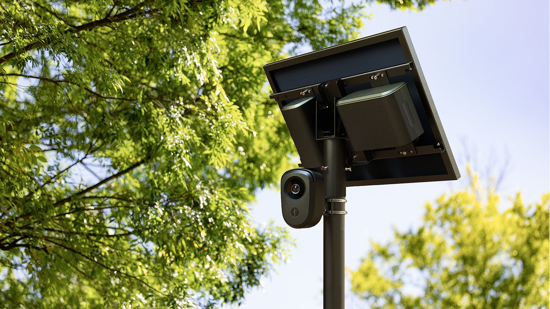 Outdoor security camera with visible lens mounted on a pole beneath a square solar panel; green trees and blue sky in a sunny park.