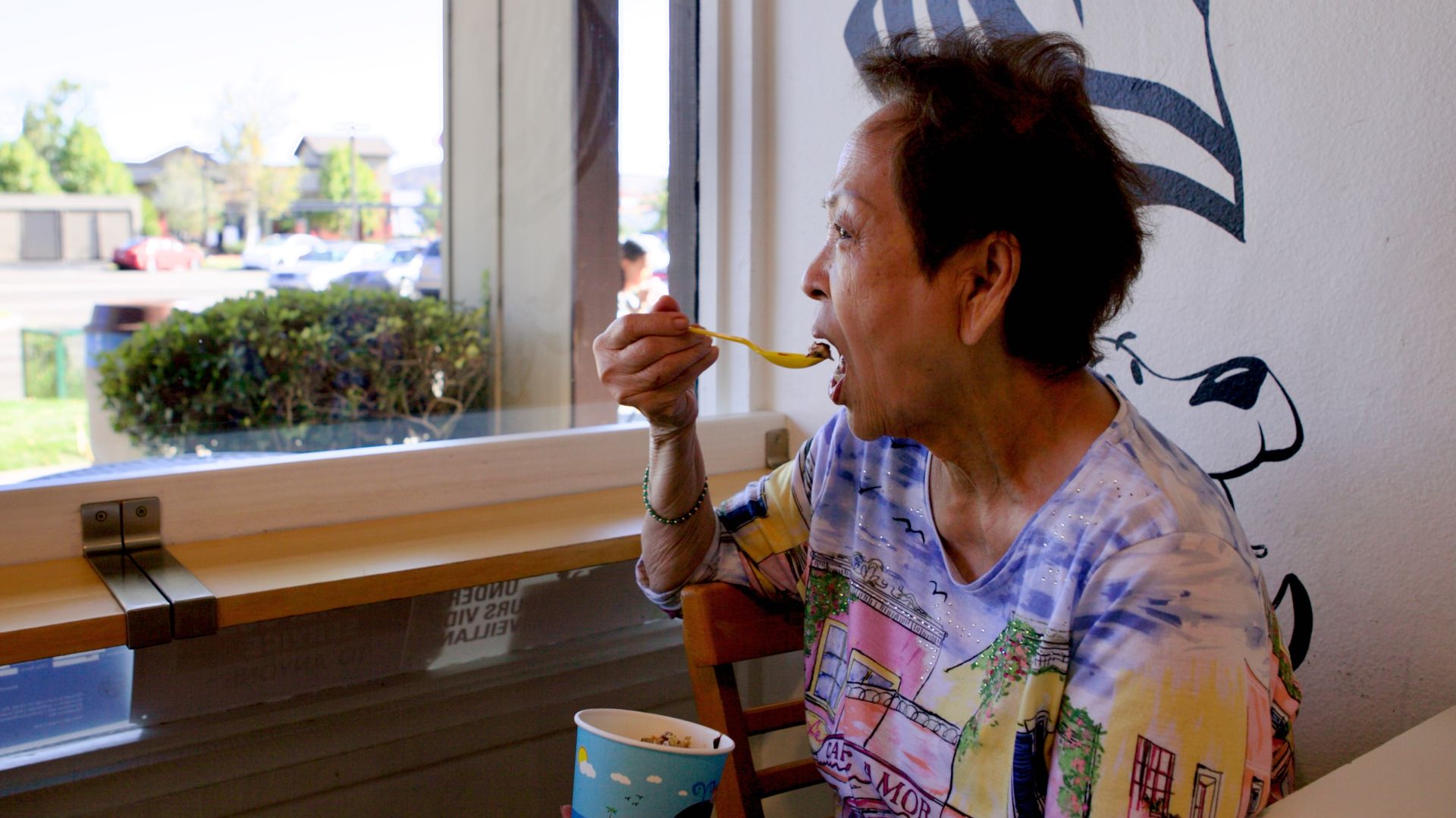 An older woman with short dark hair wearing a colorful shirt sits at a cafe table, eating with a yellow spoon. A blue mural on the wall and a window showing parked cars outside.