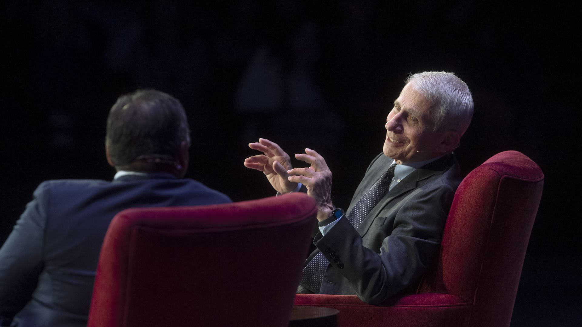 A picture of two people (one of them being Anthony Fauci smiling with his hands in front of him) sitting in big red chairs