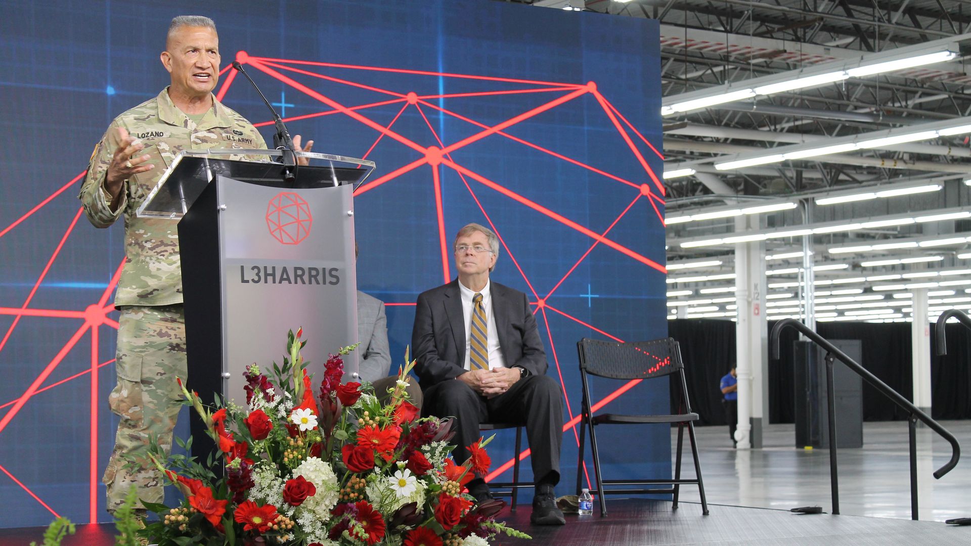 A man in U.S. Army camo uniform speaks at an L3Harris podium with a large digital network graphic behind him. A man in a suit and tie sits beside the stage with flowers in front.