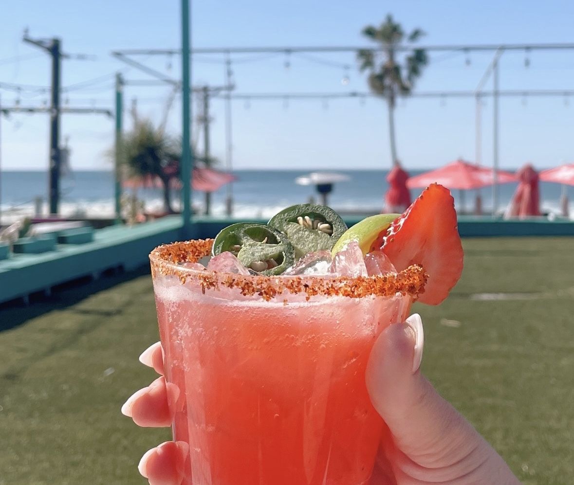 A pink margarita in a glass topped with jalepenos, a strawberry, a lime wedge and a Tajin rim held in the air at a rooftop bar overlooking the beach.