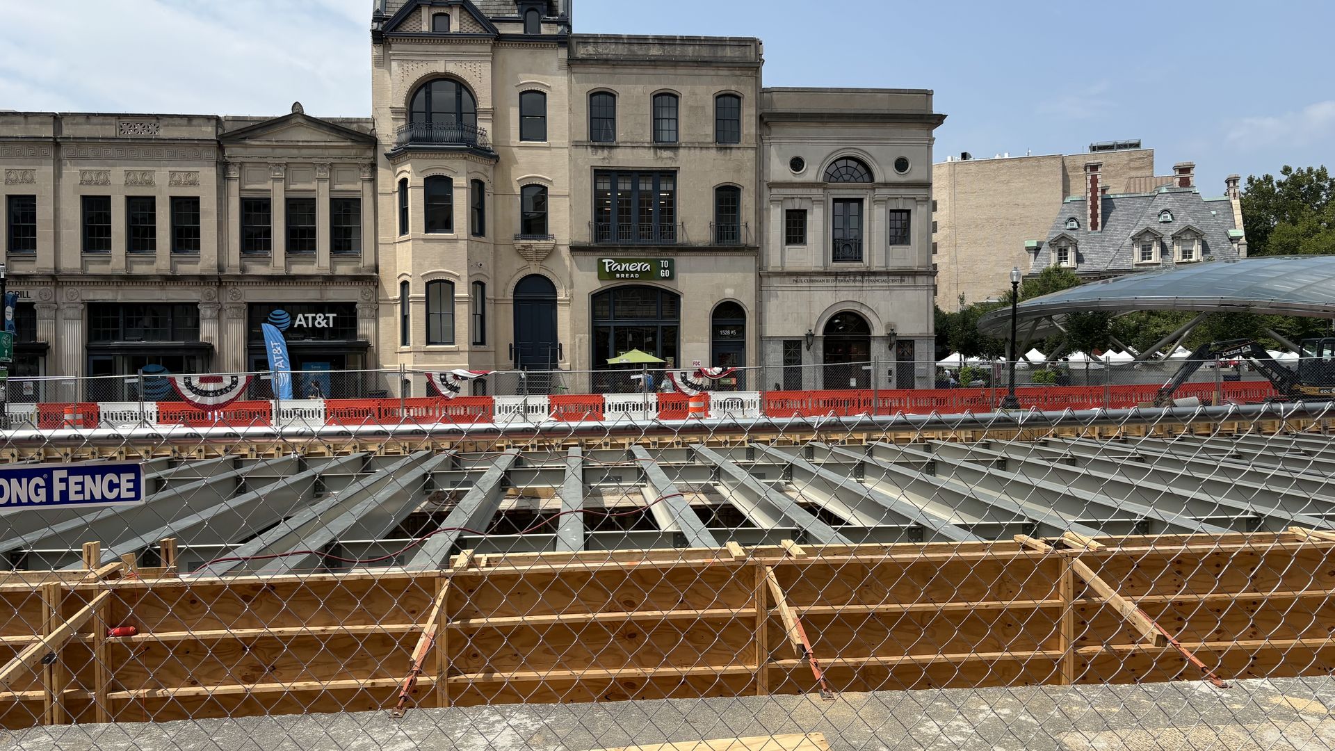 Historic beige storefronts line a city street under a blue sky. In front is a construction site with wooden framing, steel beams, and chain-link fencing; AT&T and Panera Bread signs visible.