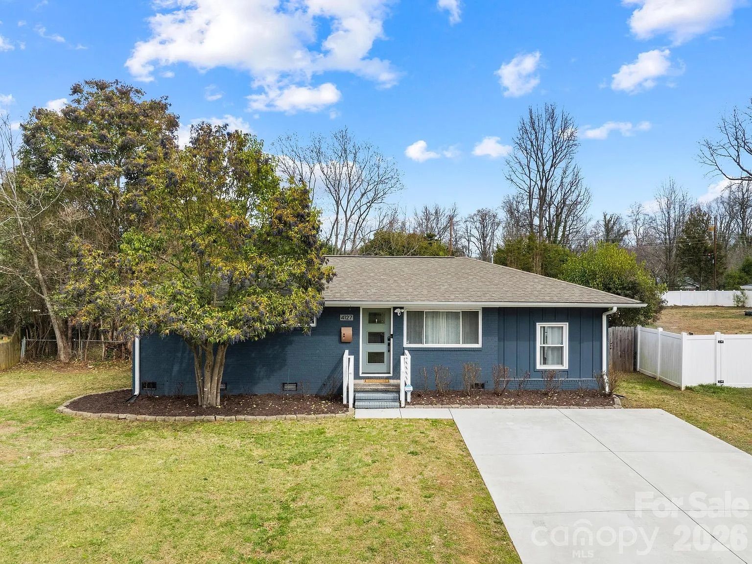 Single-story blue house with gray roof, a large tree on the left, concrete driveway on the right, green lawn, and a sunny sky with scattered clouds.