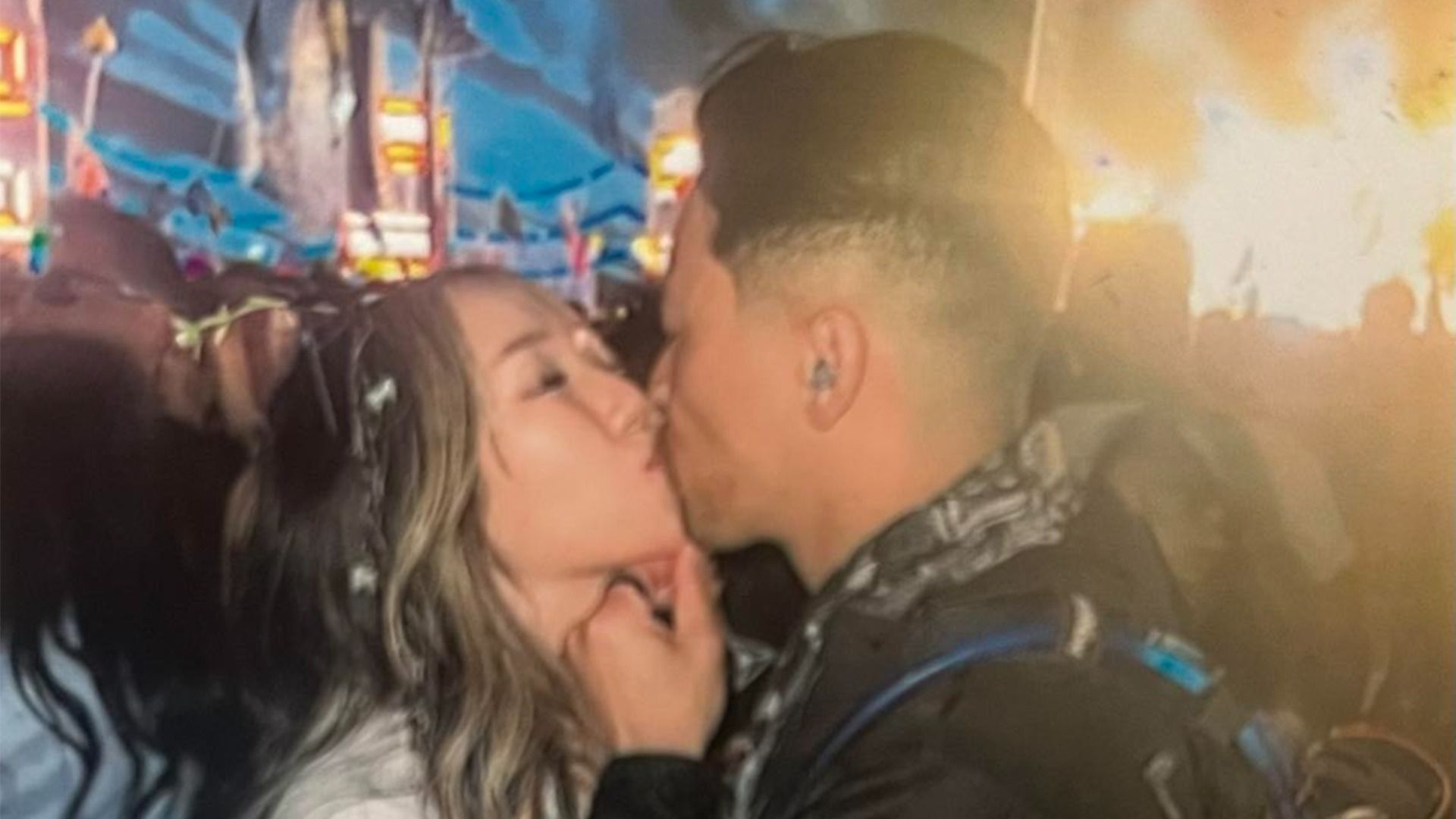 A photo of a couple kissing at an electronic music festival with lights blazing in the background. 