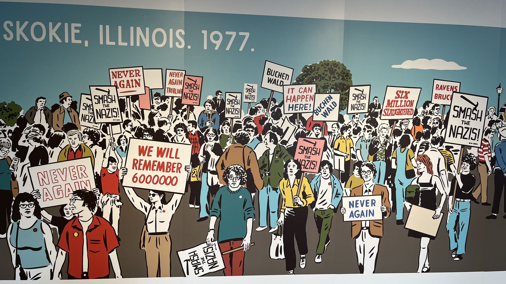 Photo of a mural showing a 1977 protest in Skokie, Illinois, with diverse people holding signs saying "Smash the Nazis!", "Never Again", "We Will Remember 6000000", and "It Can Happen Here!" against a blue sky.