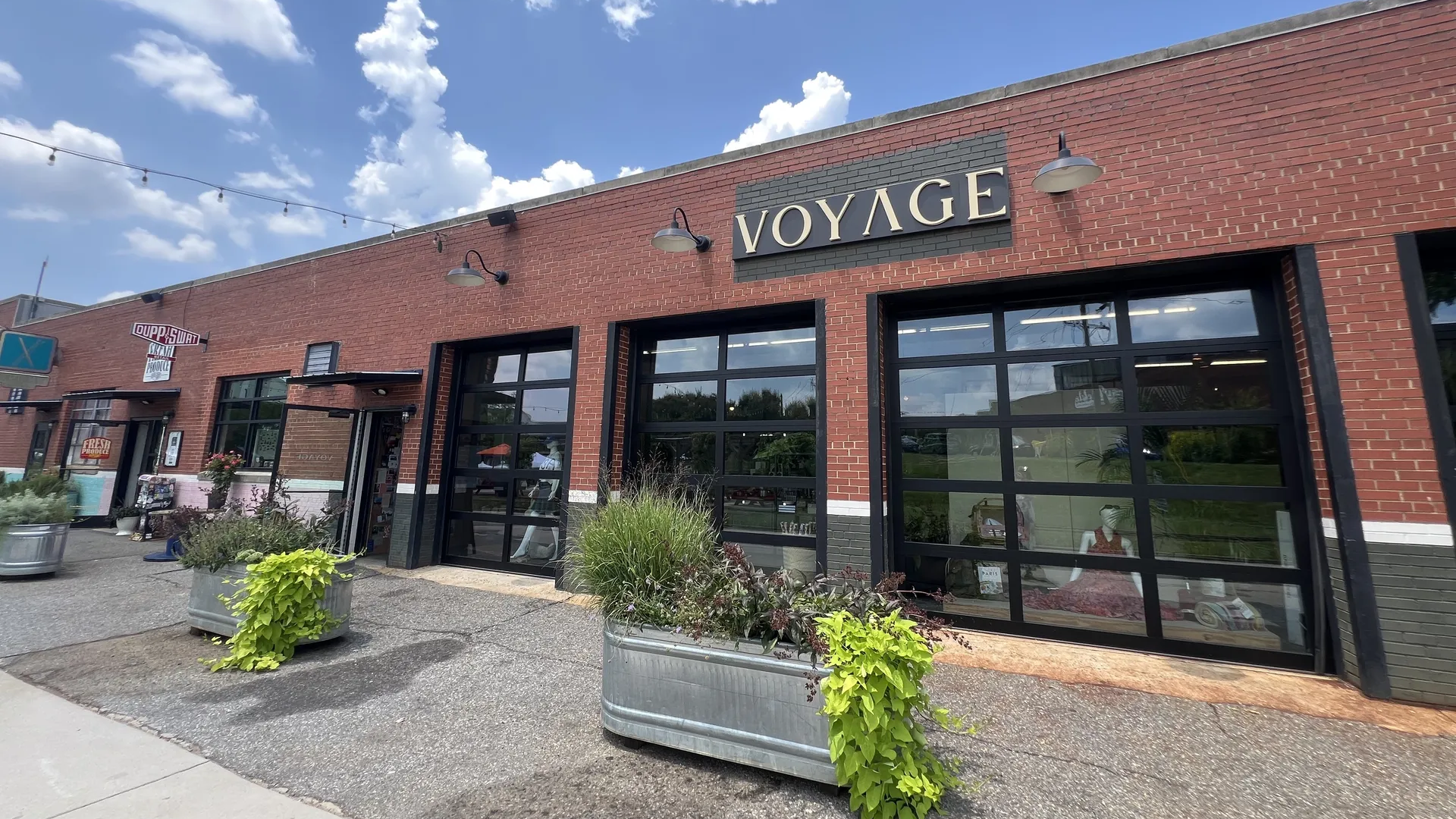 Exterior of a red brick building with a black and gold sign reading "VOYAGE," featuring large black framed windows and planters with green plants under a blue sky with white clouds.