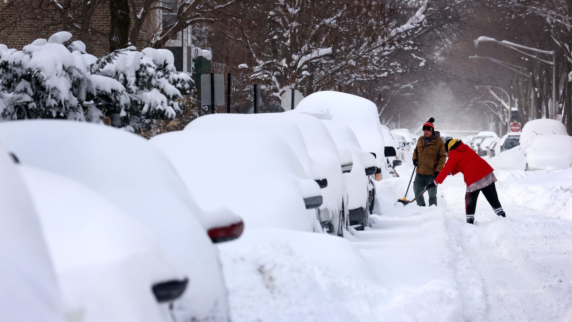 Photo of two people shoveling snow next to a line of cars