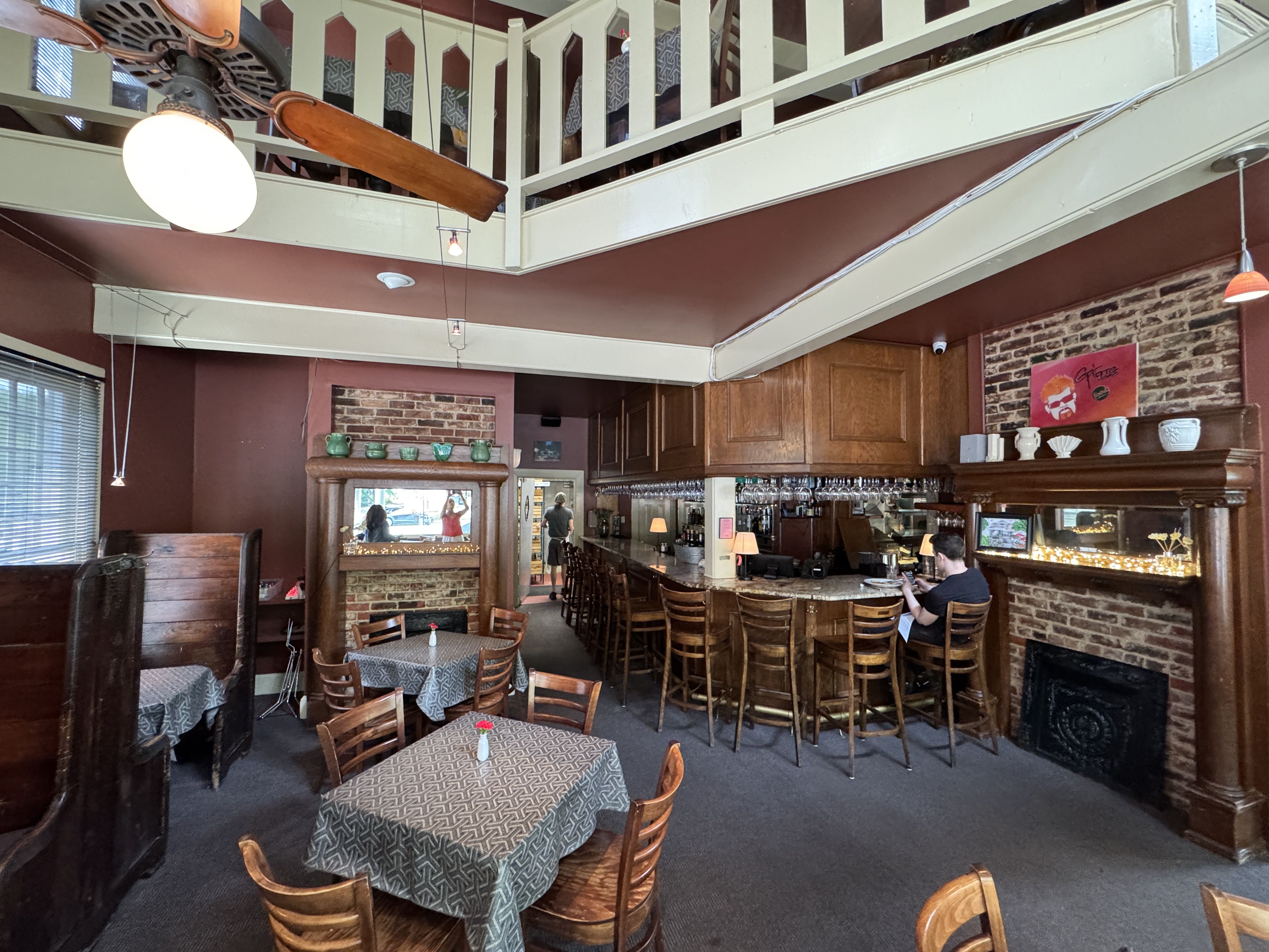 A cozy cafe interior with a warm color scheme. The room features several wooden tables and chairs covered with patterned tablecloths. In the background, there's a bar with several stools and a person sitting at the counter.