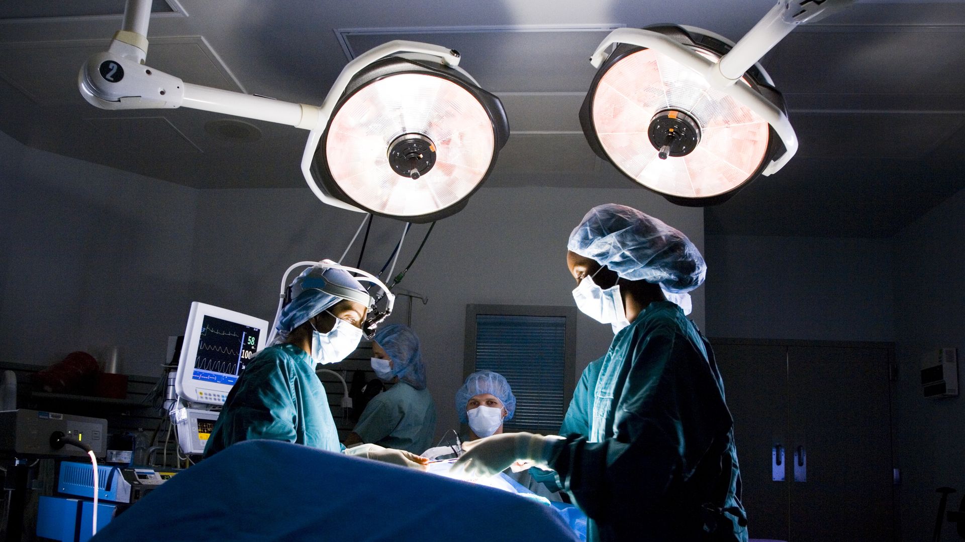 In this image, three surgeons stand under bright lights in a dark operating room. 