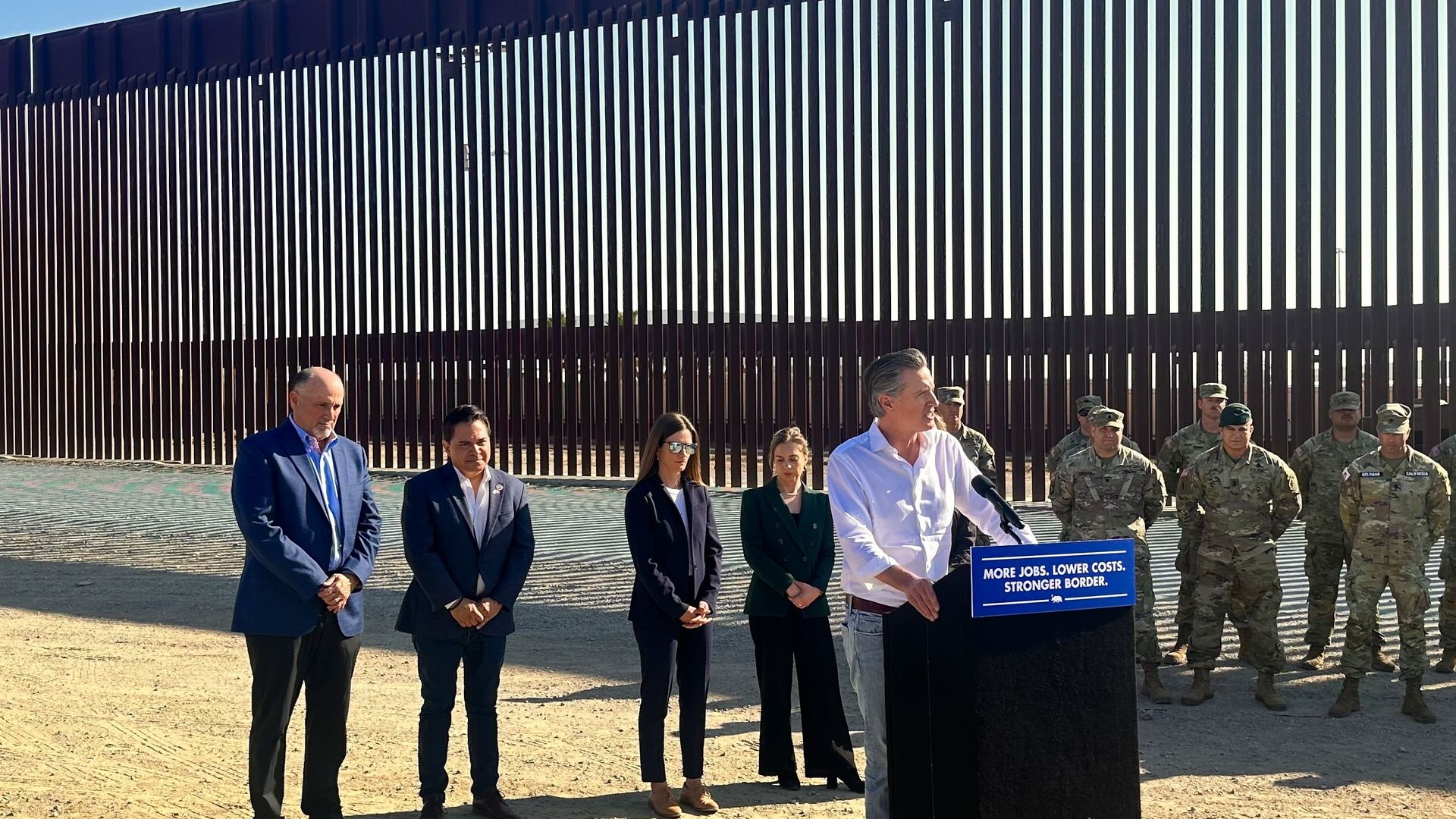 California Gov. Gavin Newesom behind a lectern at the border wall with members of the California National Guard and other officials.