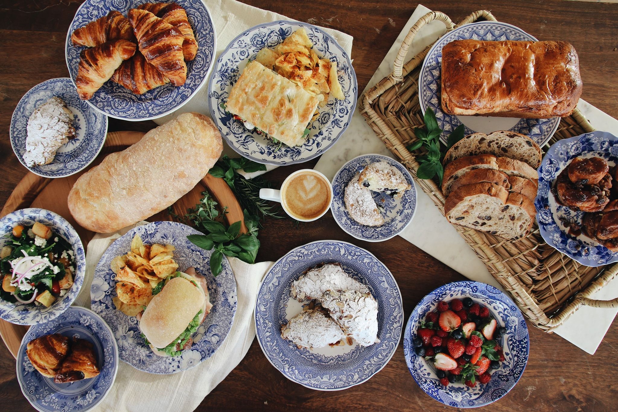 Variety of breads, pastries with powdered sugar, sandwiches with chips, fruit salad, salad with croutons, and a latte on blue and white floral plates on a wooden table.