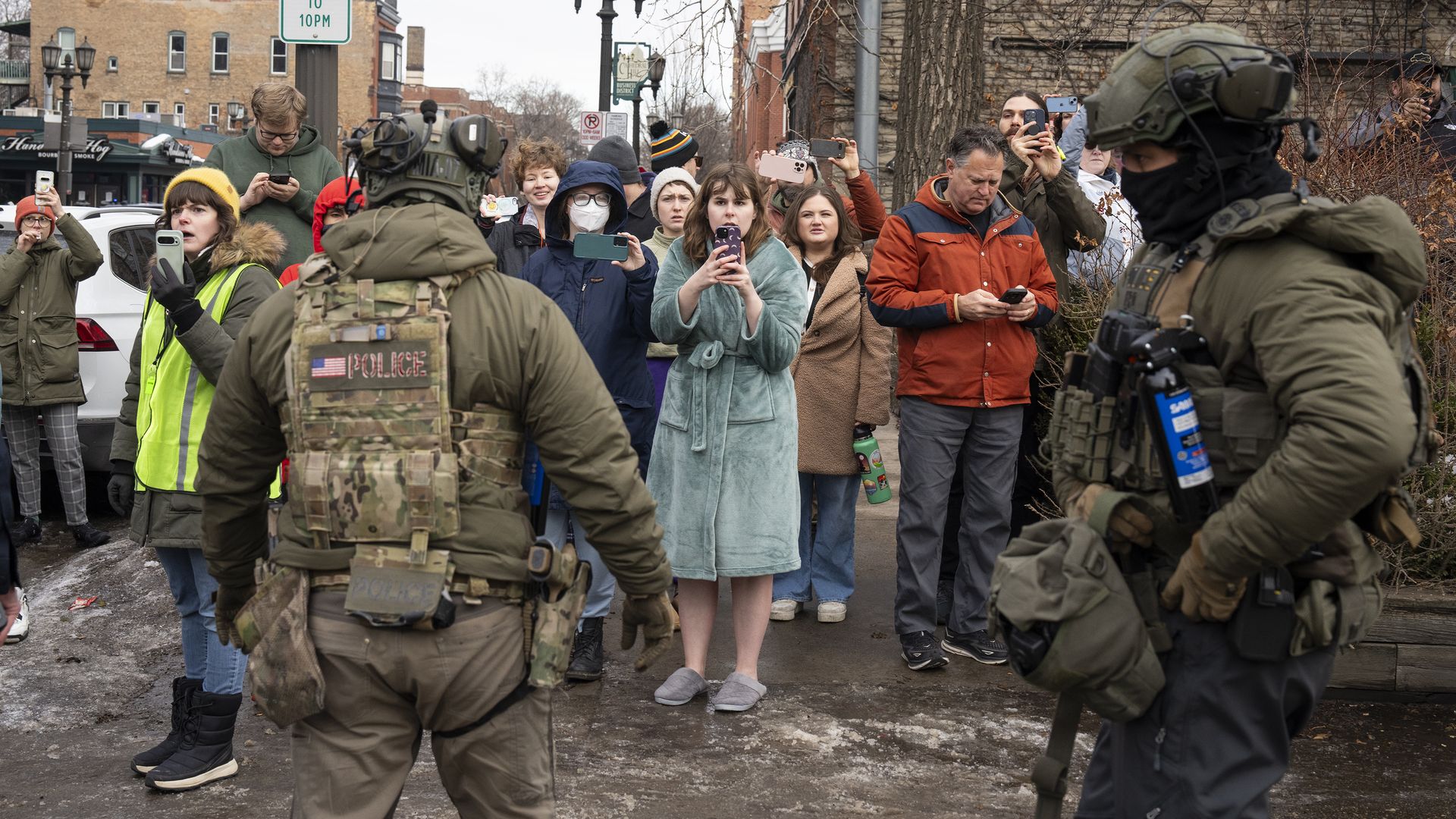 Two heavily armed police in tactical gear face a crowd of onlookers on a city street. People wearing winter clothes record the scene with phones. A 2-hour parking sign is visible.