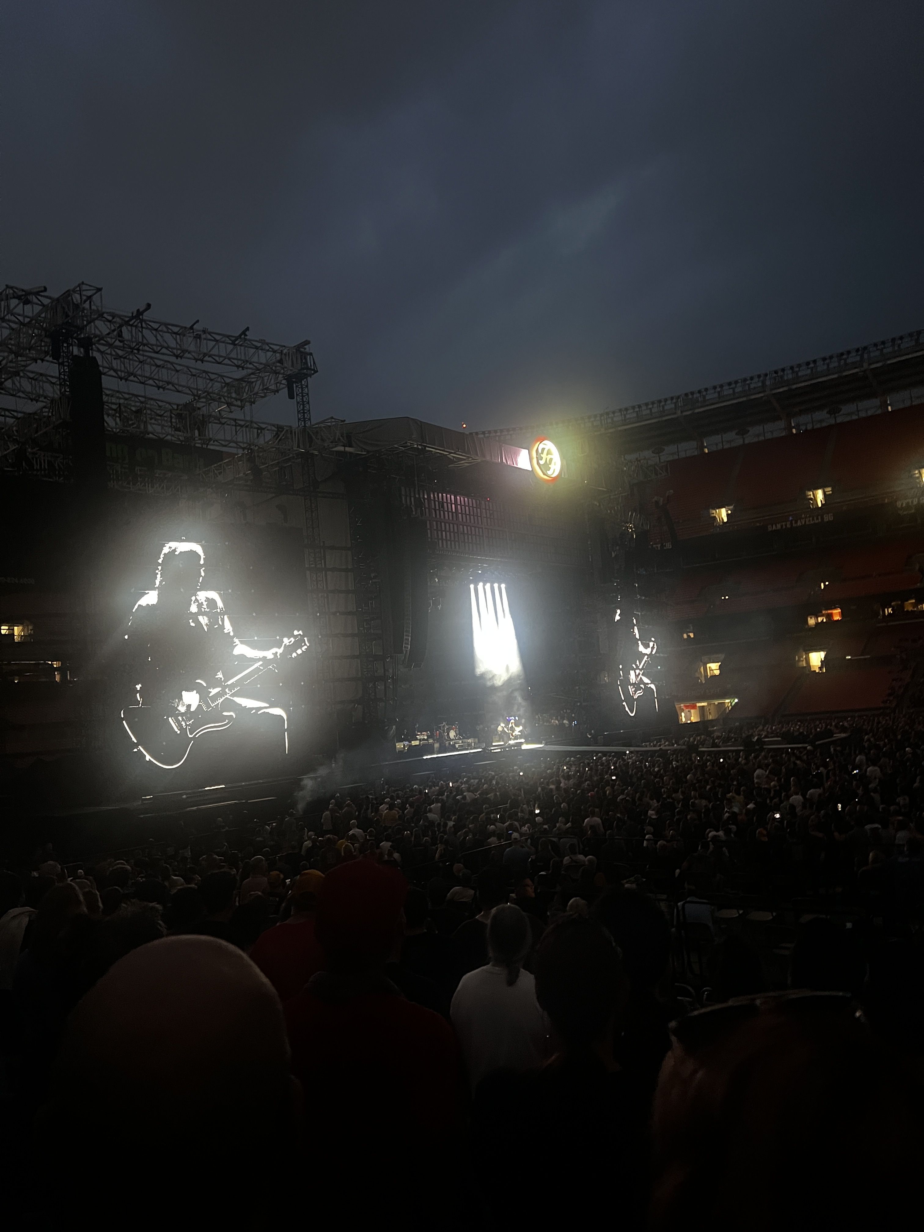 Nighttime stadium concert with a large stage; bright white guitar outline on the left screen, a tall central LED panel, a glowing circular emblem above the stage, and a dense crowd in front.