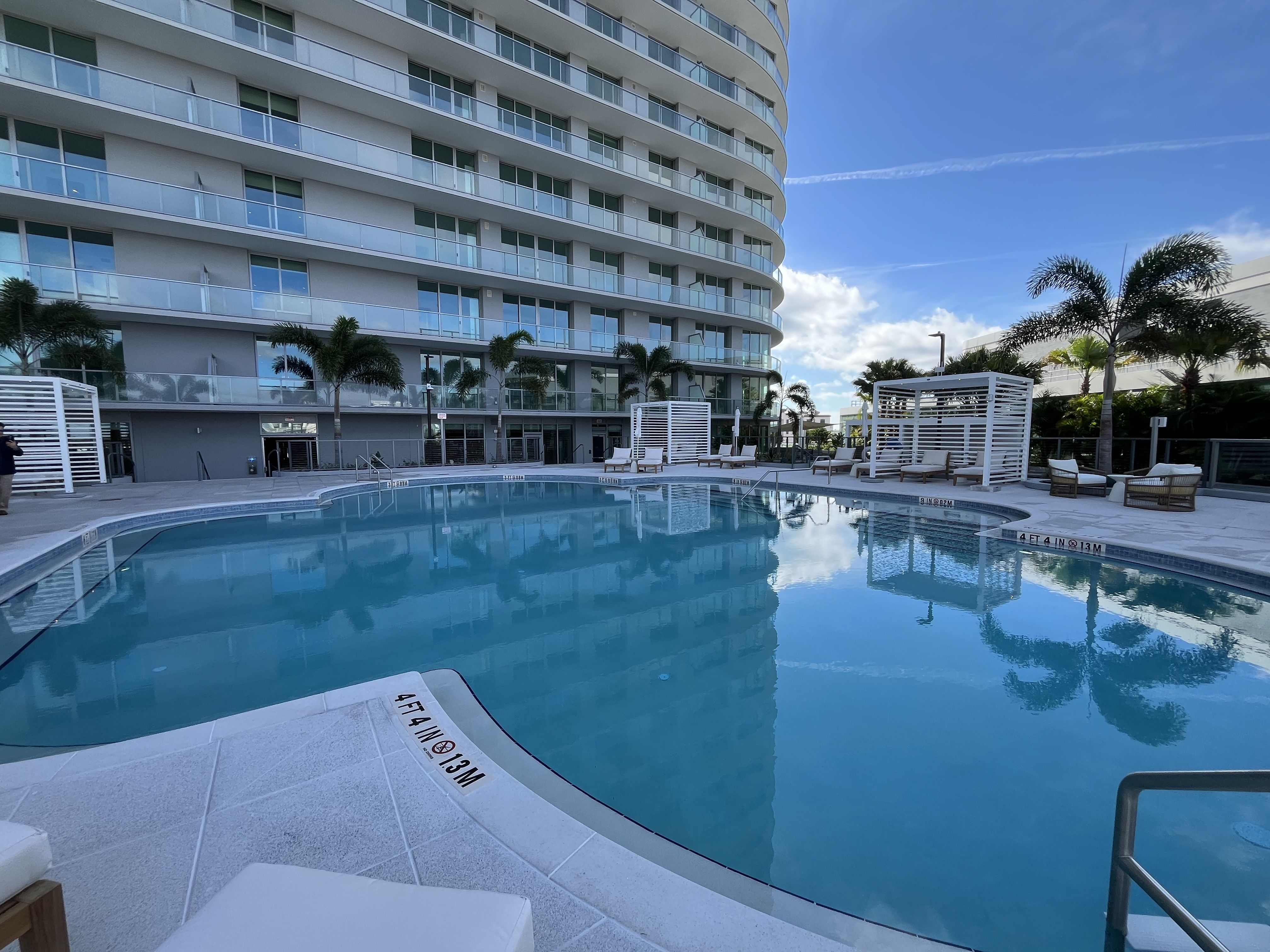 Clear blue swimming pool surrounded by white lounge chairs, palm trees, and modern curved glass building under a bright blue sky with scattered clouds.