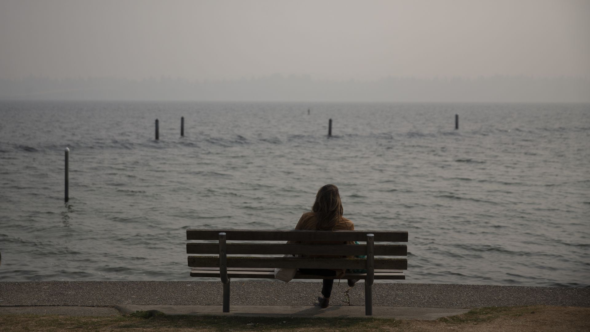 A person sits on a bench looking at the water against a smoky sky. 