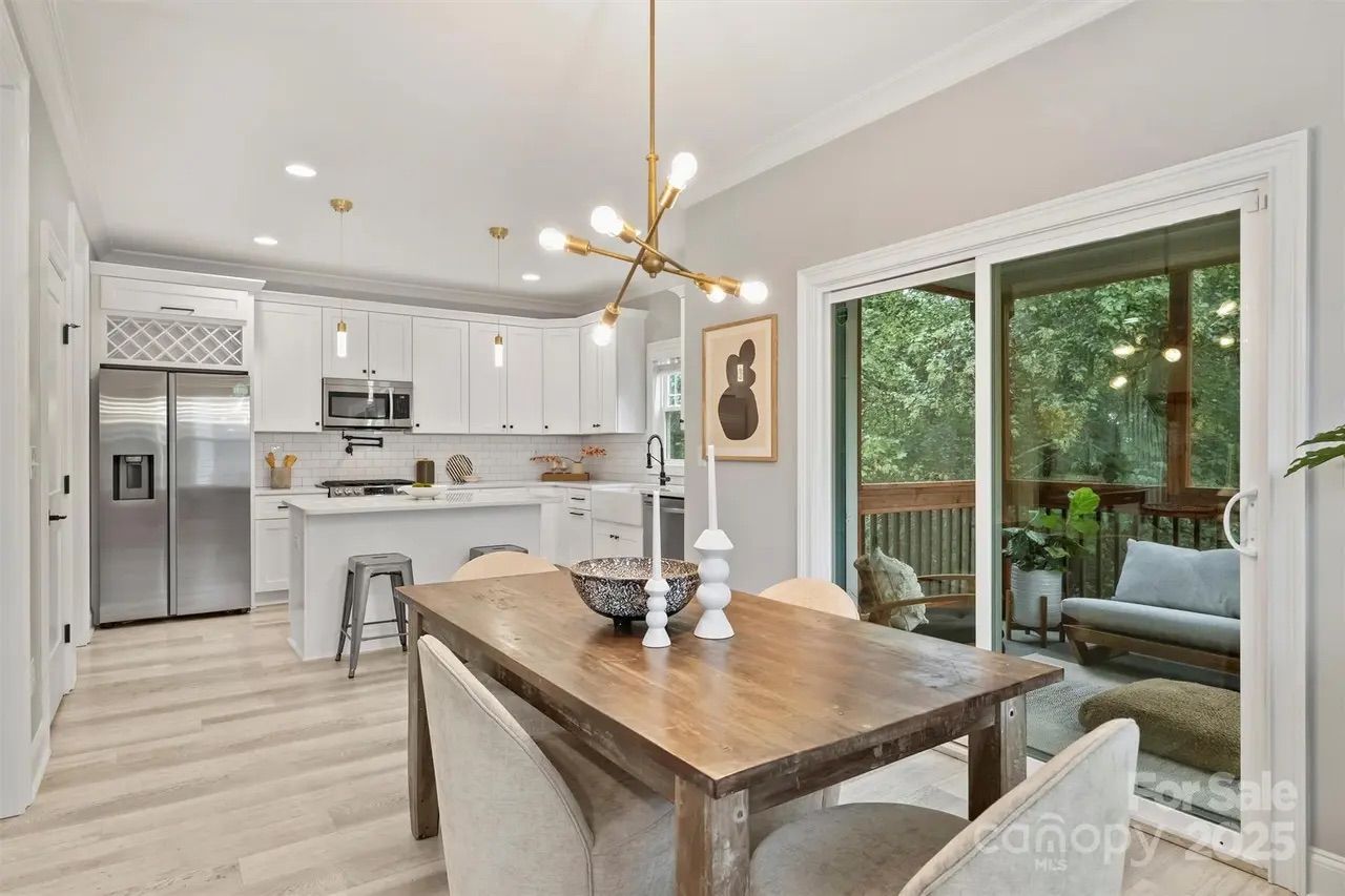 Bright modern kitchen and dining area with white cabinets, stainless steel appliances, wooden dining table with chairs, gold light fixture, and sliding glass doors leading to a green outdoor deck.