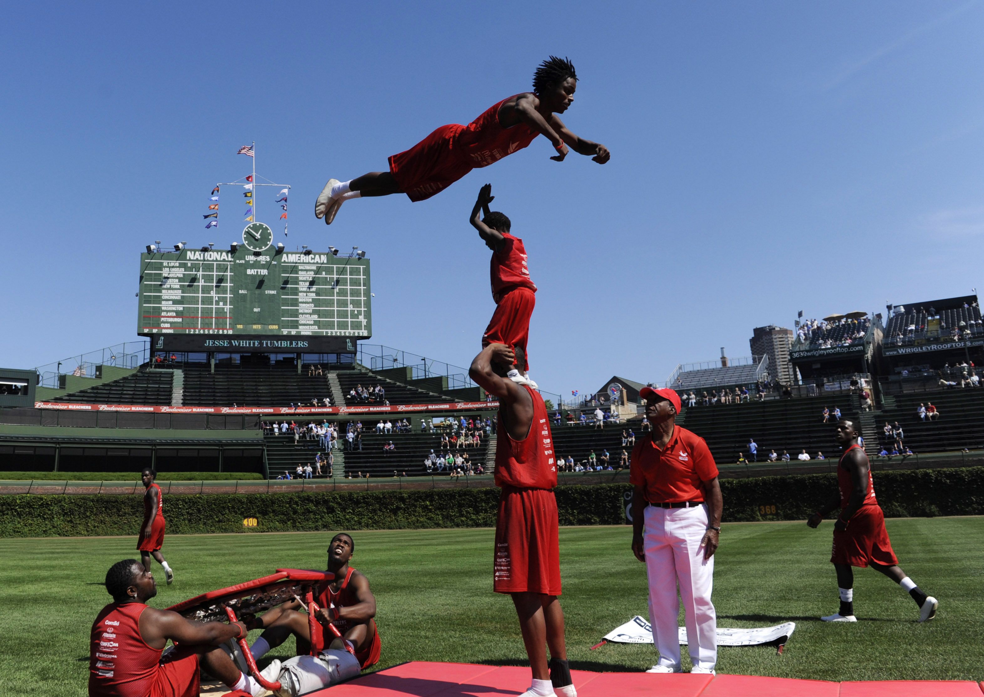 Performers in red outfits execute a tumbling stunt on a baseball field with a large green scoreboard in the background under a clear blue sky.