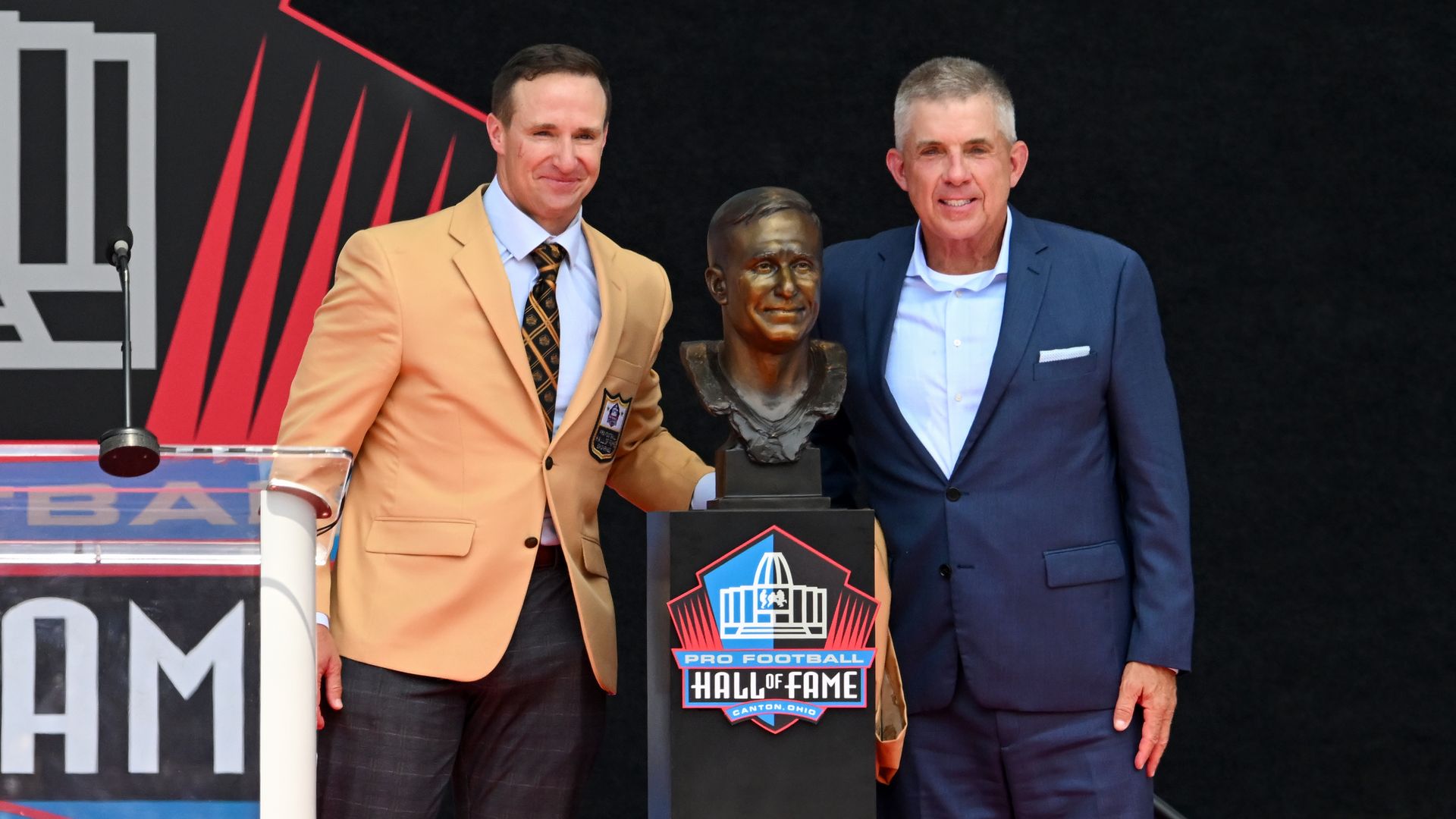 Drew Brees, wearing a gold jacket and tie, stands with Sean Payton next to a gold bust sculpture of Brees.