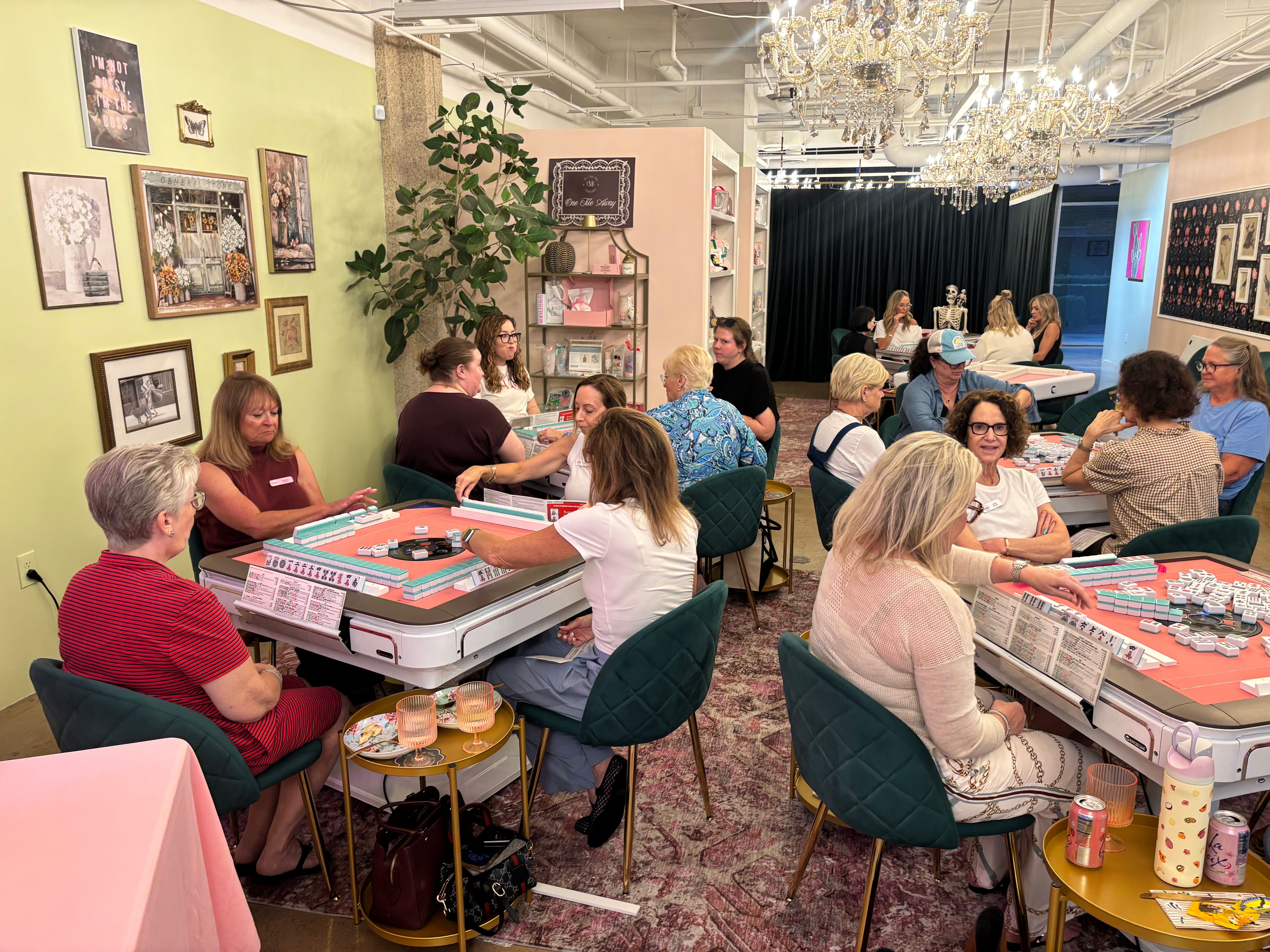 A bright, busy interior with players seated at pink card tables playing Mahjong; green cushioned chairs, wall art, a tall plant, shelves, and a crystal chandelier overhead.